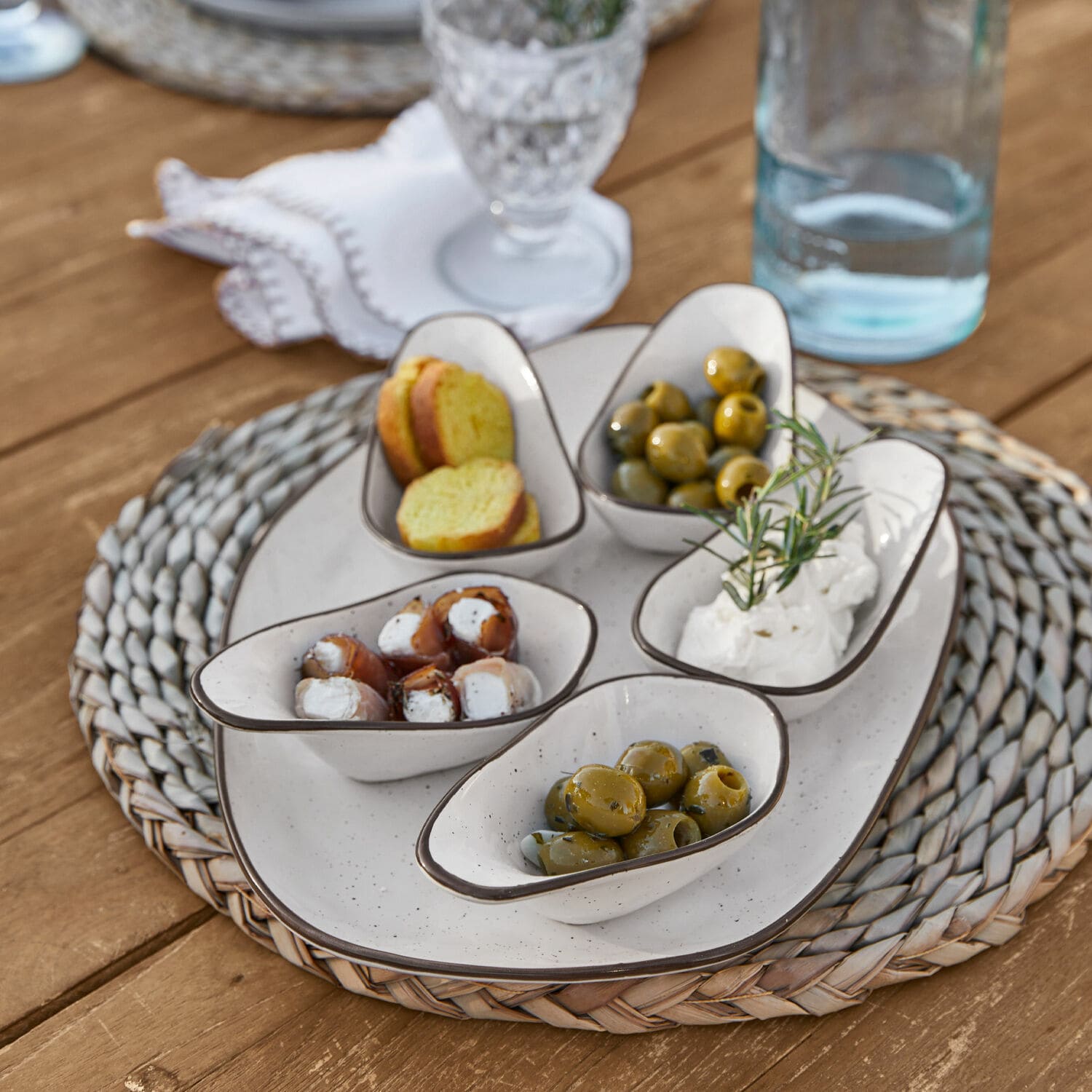 Set de 5 cuencos y plato de servir Biarré Pequeños cuencos de cerámica blanca con aceitunas y queso sobre base trenzada de madera
