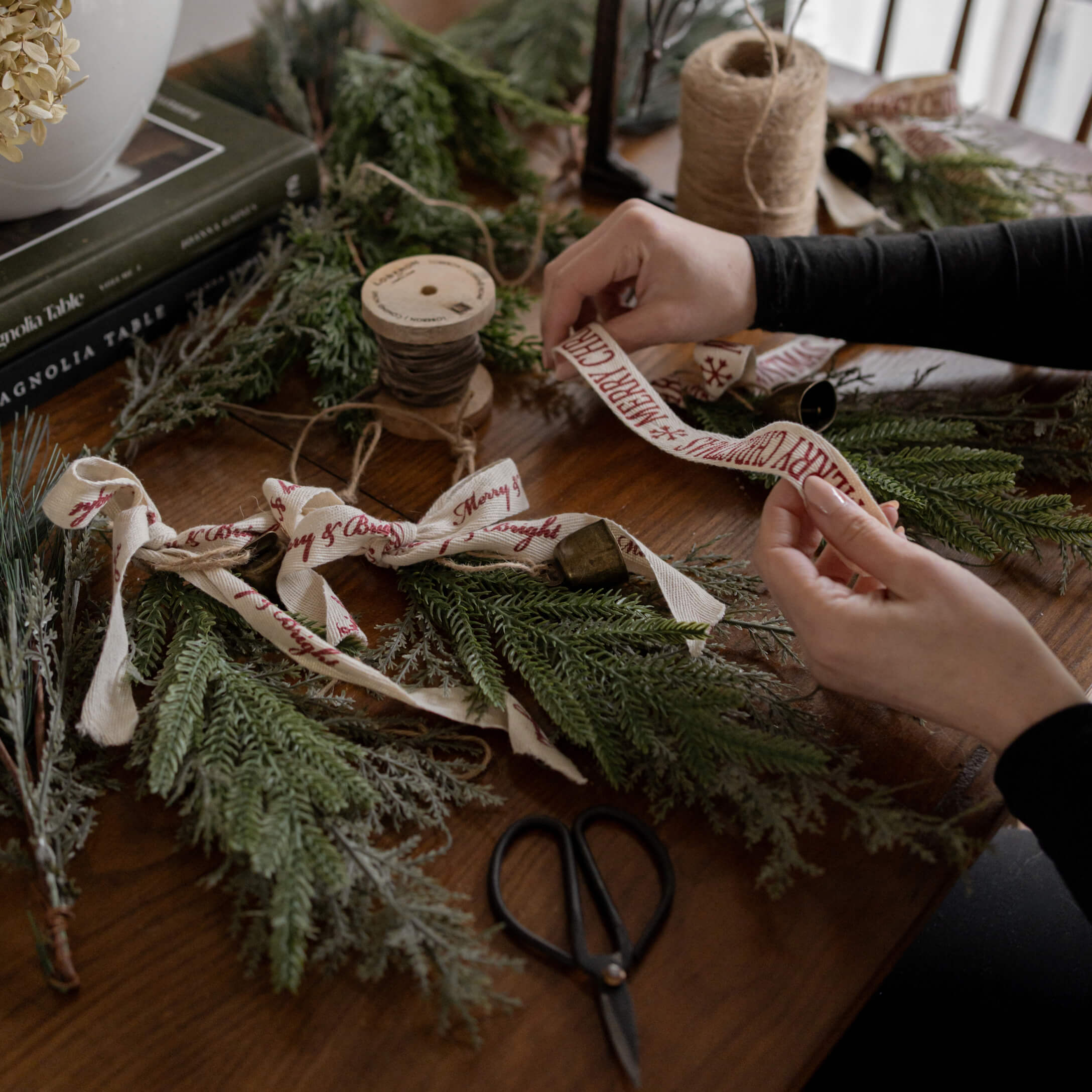 Manos decorando ramas verdes con cinta de algodón Merry Christmas sobre mesa de madera con tijeras