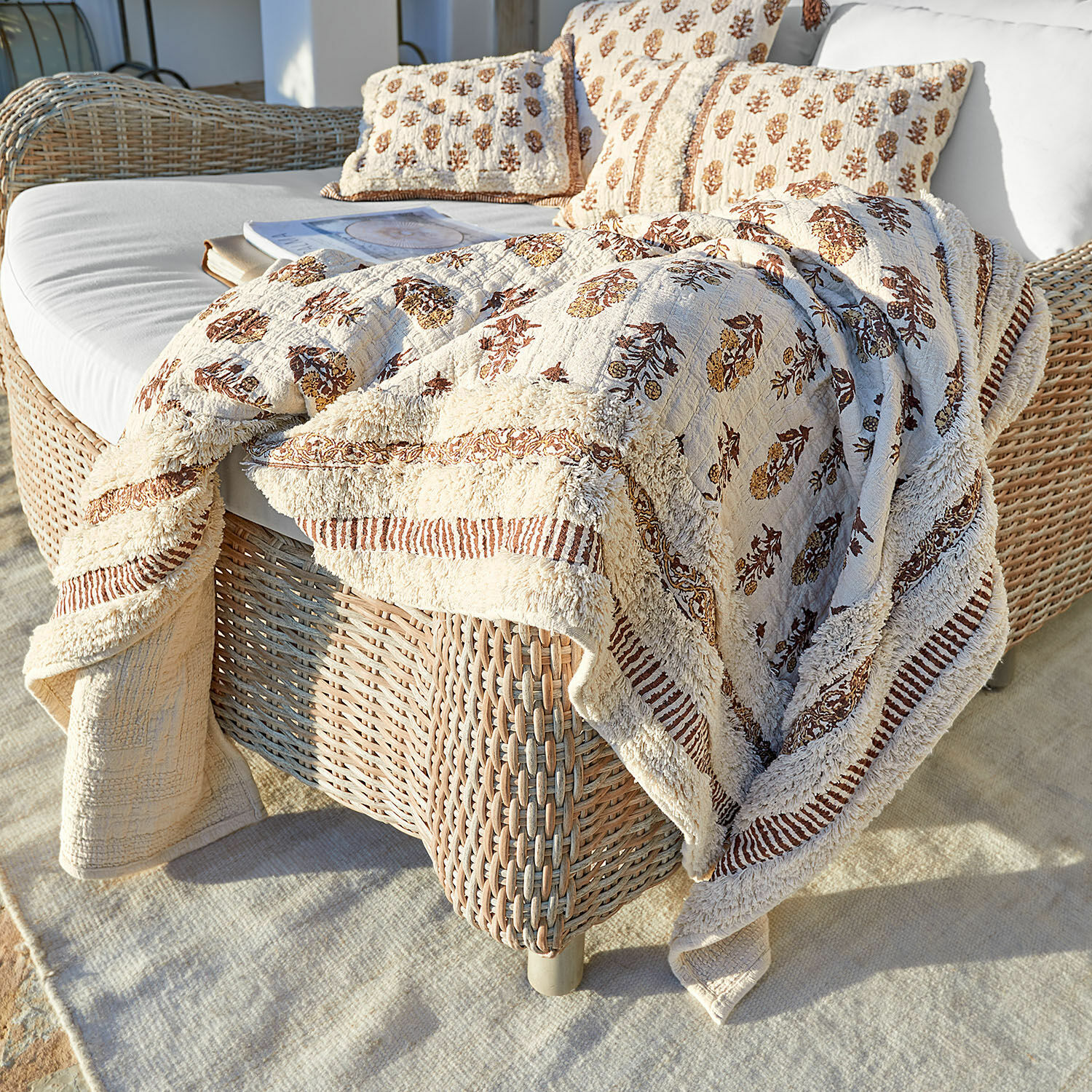 Colcha Arsalan Manta y cojines de algodón beige con diseño marrón sobre sofá de mimbre en terraza