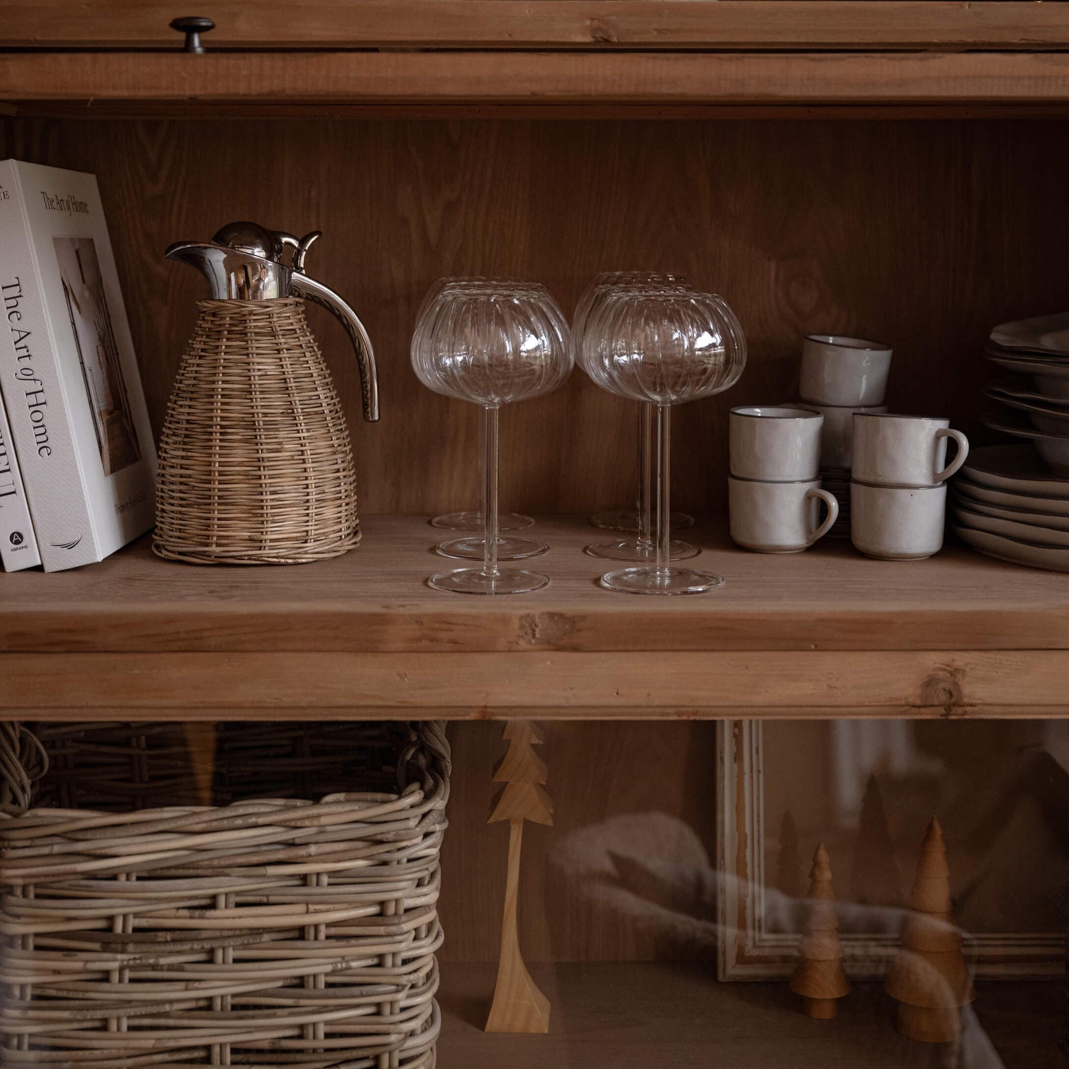 Copas de cristal con diseño acanalado en estante de madera con jarra de mimbre y tazas blancas apiladas