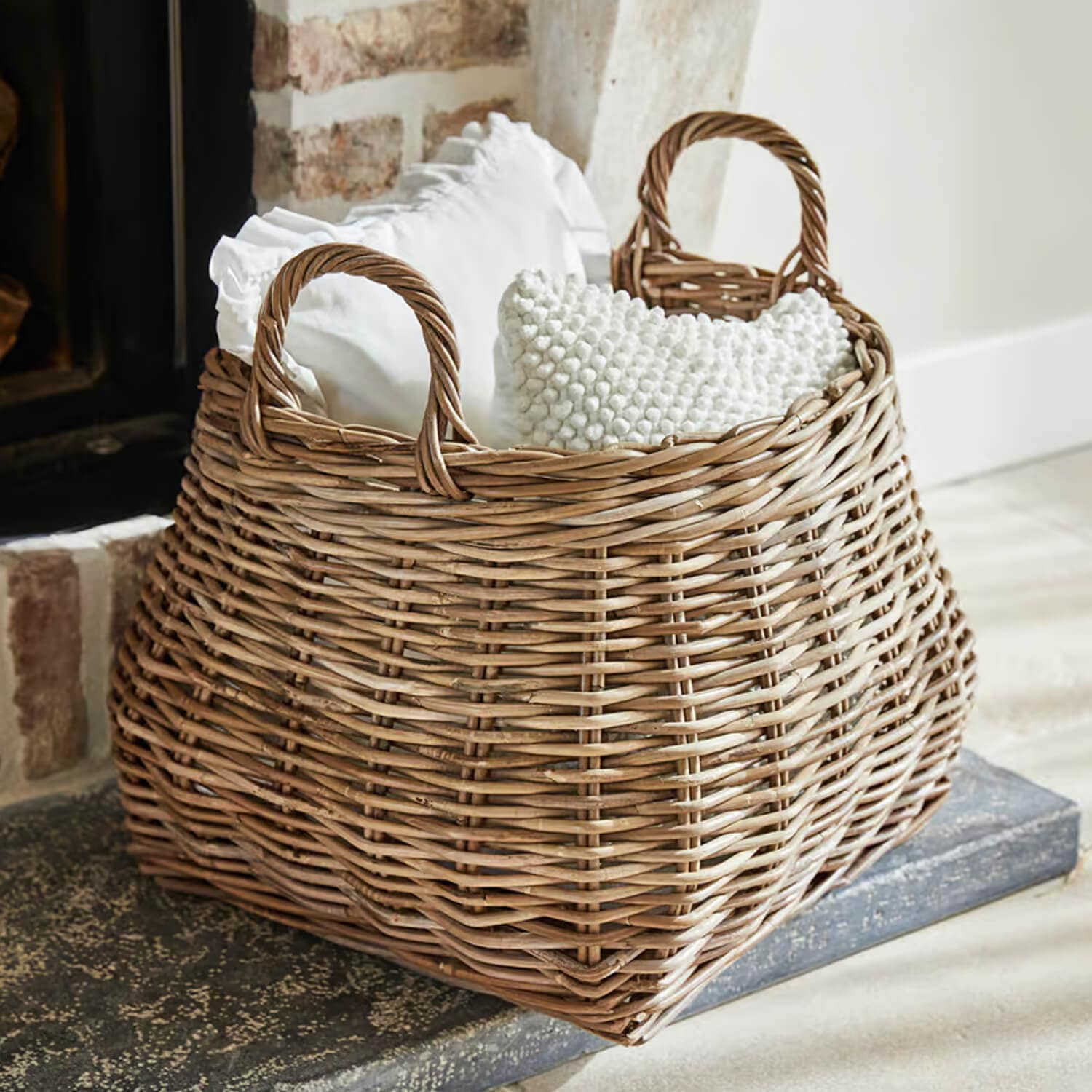 Cesta de mimbre natural con asas, rellena de cojines blancos, ideal para almacenamiento y decoración