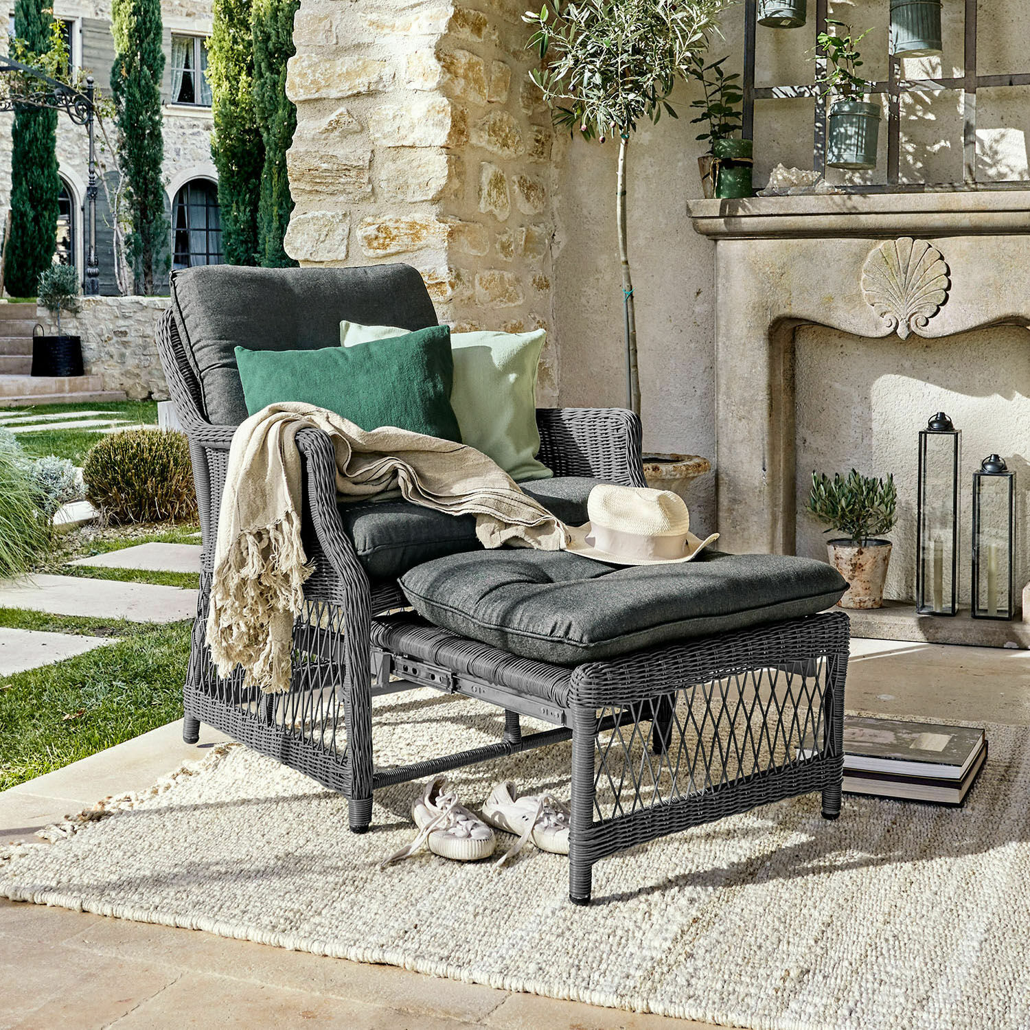 Sillón de ratán gris con reposapiés y cojines gris oscuro, decorado con cojines verdes en una terraza