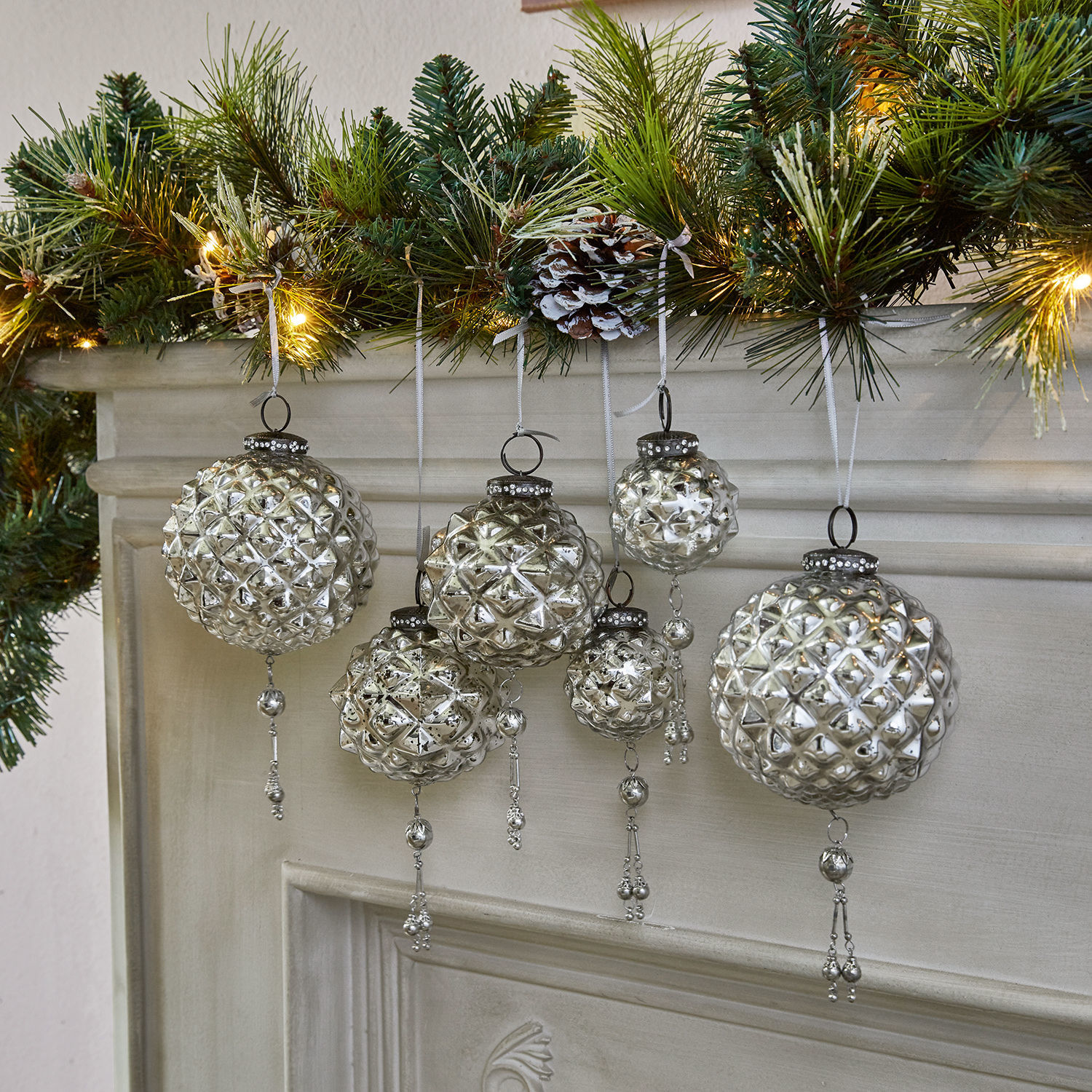 Set de 6 adornos navideños Murron Bolas de Navidad de vidrio plateado con adornos colgantes frente a chimenea y ramas verdes con luces