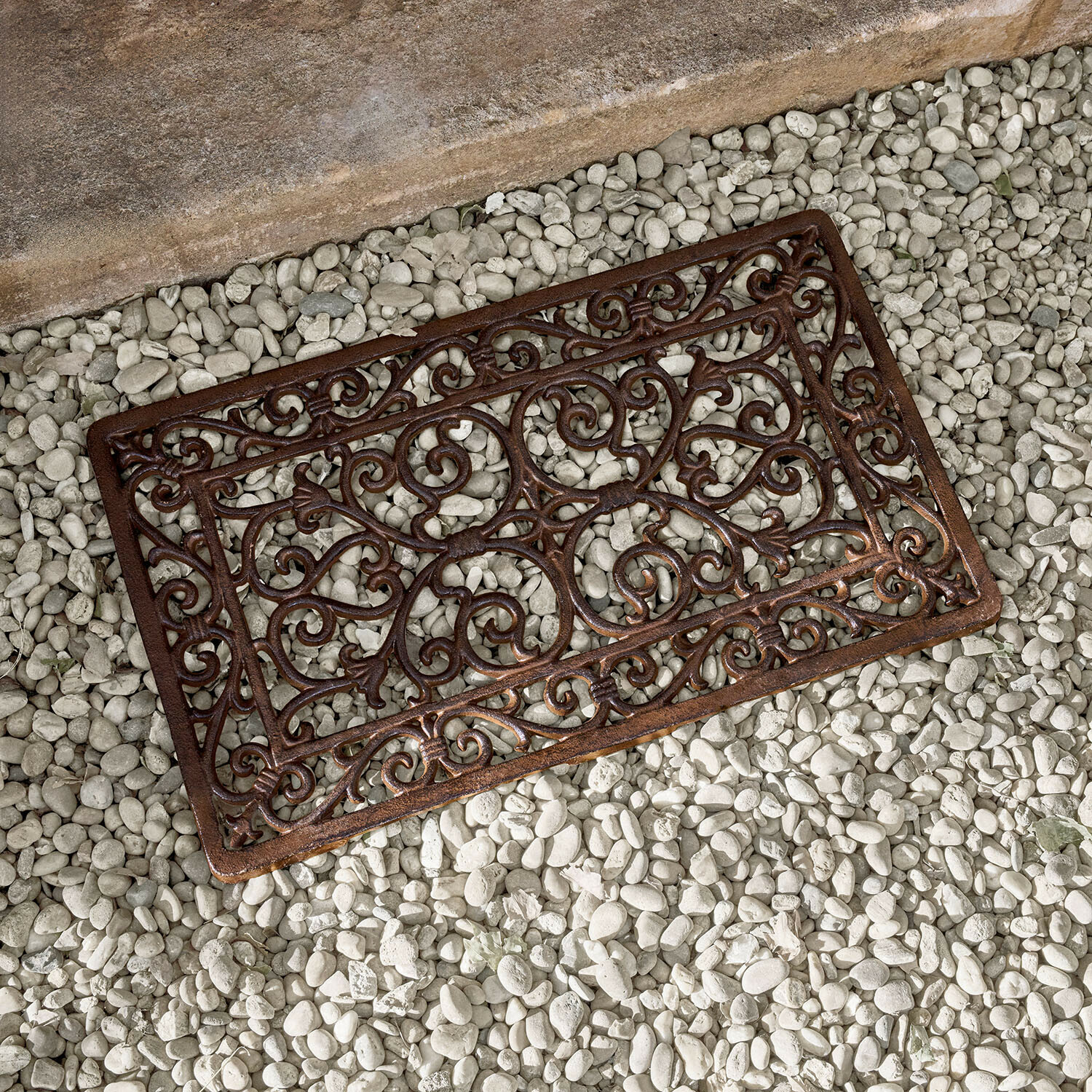 Felpudo metálico marrón con diseño ornamental sobre piedras blancas, alfombrilla de hierro fundido