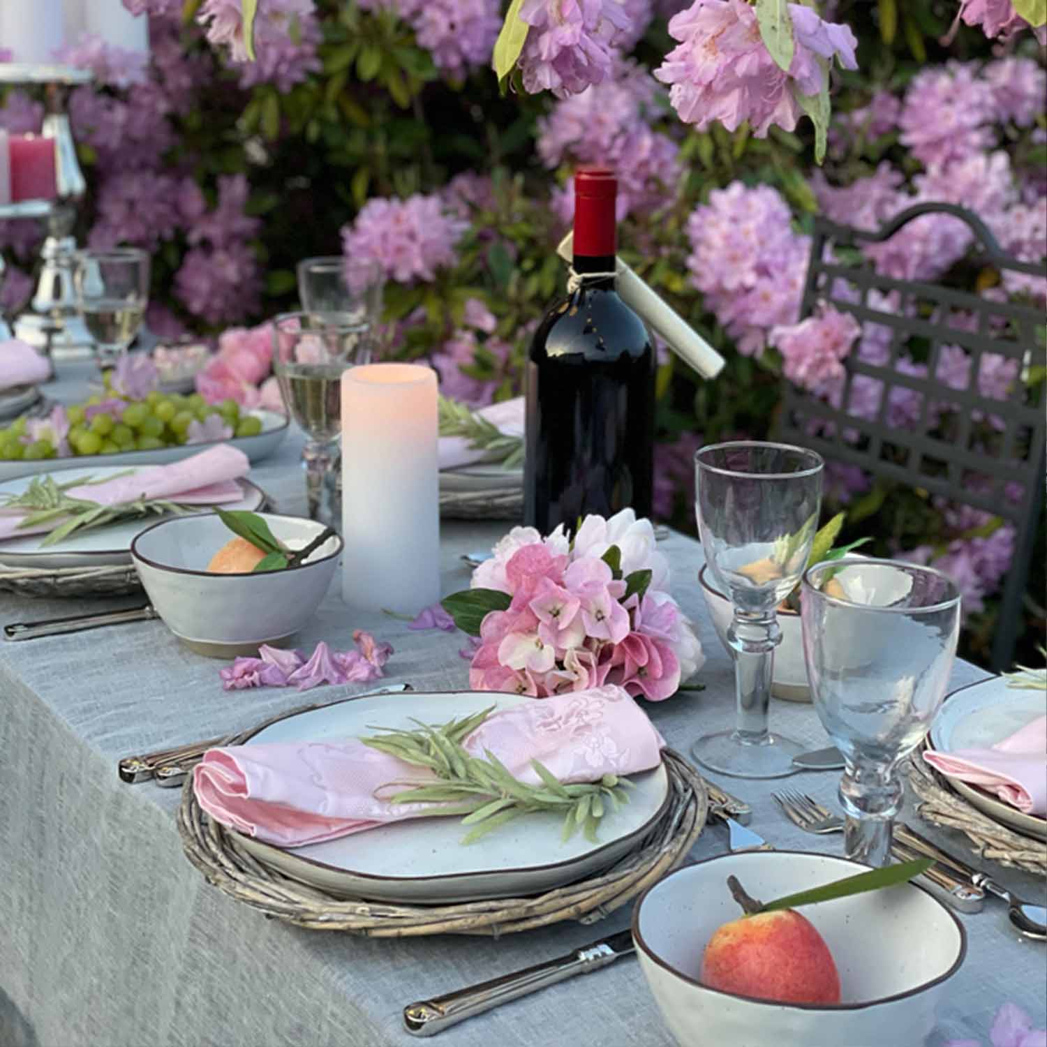 Mesa elegantemente puesta con vajilla blanca, servilletas rosas y botella de vino tinto en jardín florido
