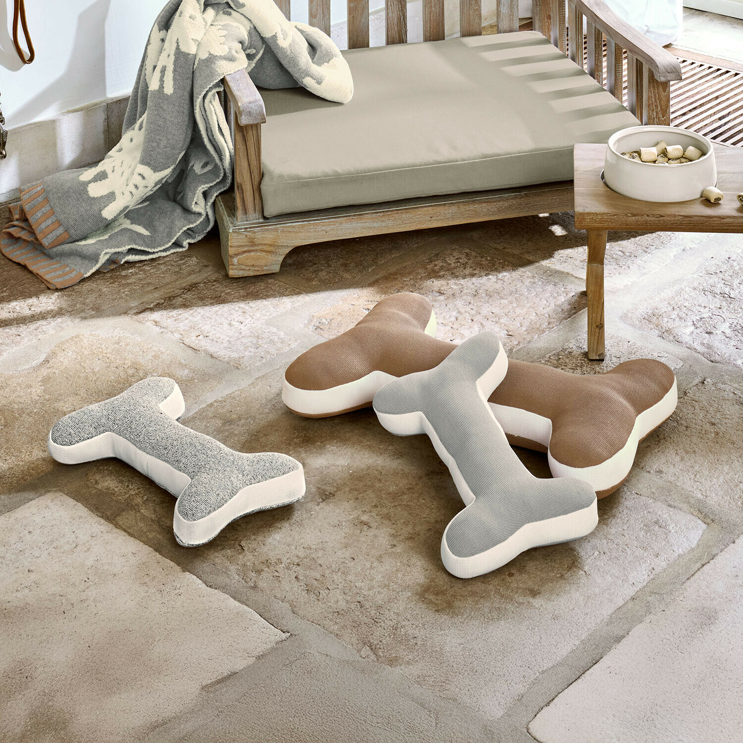 Tres cojines con forma de hueso en gris y marrón sobre suelo de piedra junto a un banco de madera con cojín beige