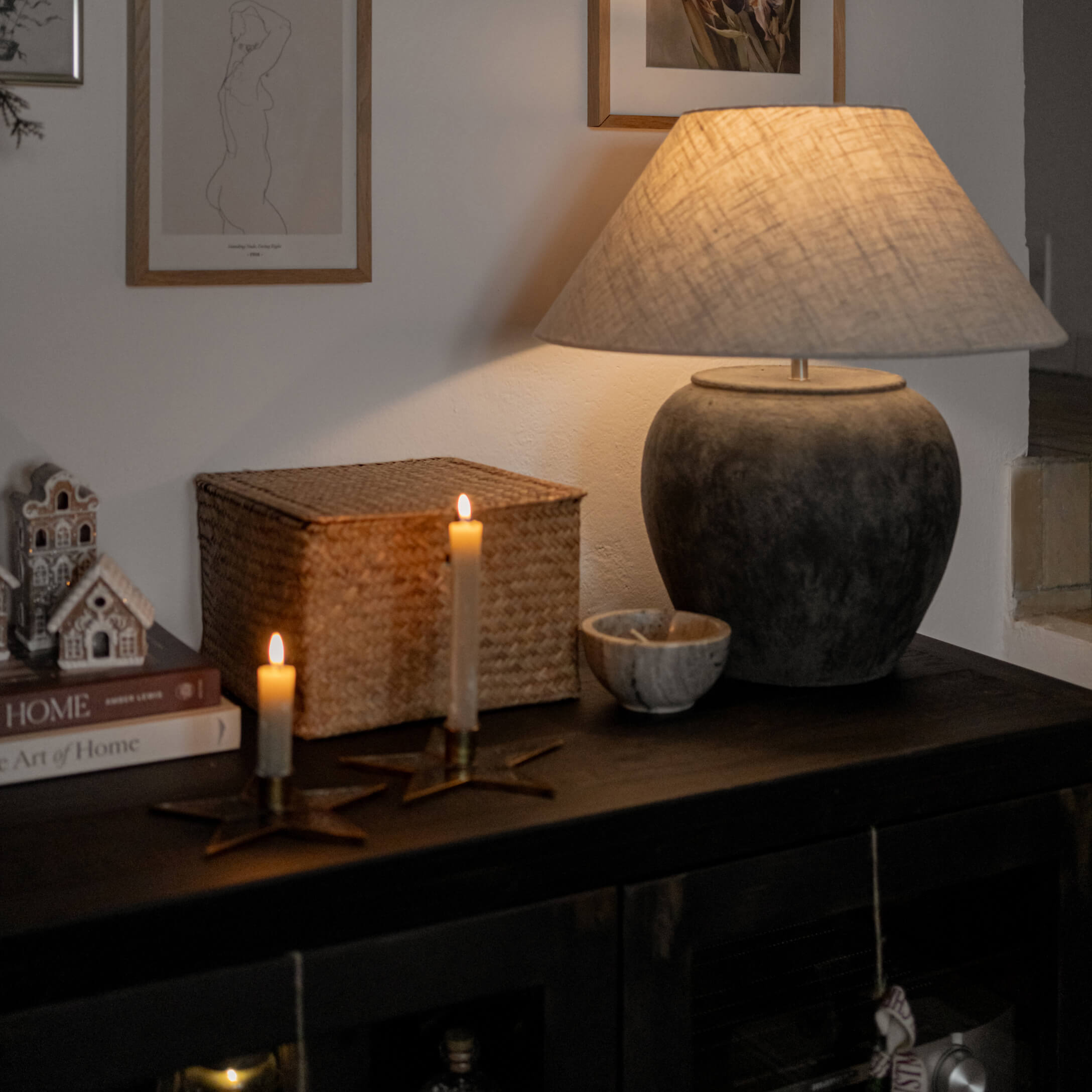 Lámpara de mesa de cerámica gris oscuro con pantalla beige sobre mueble oscuro con velas y decoraciones