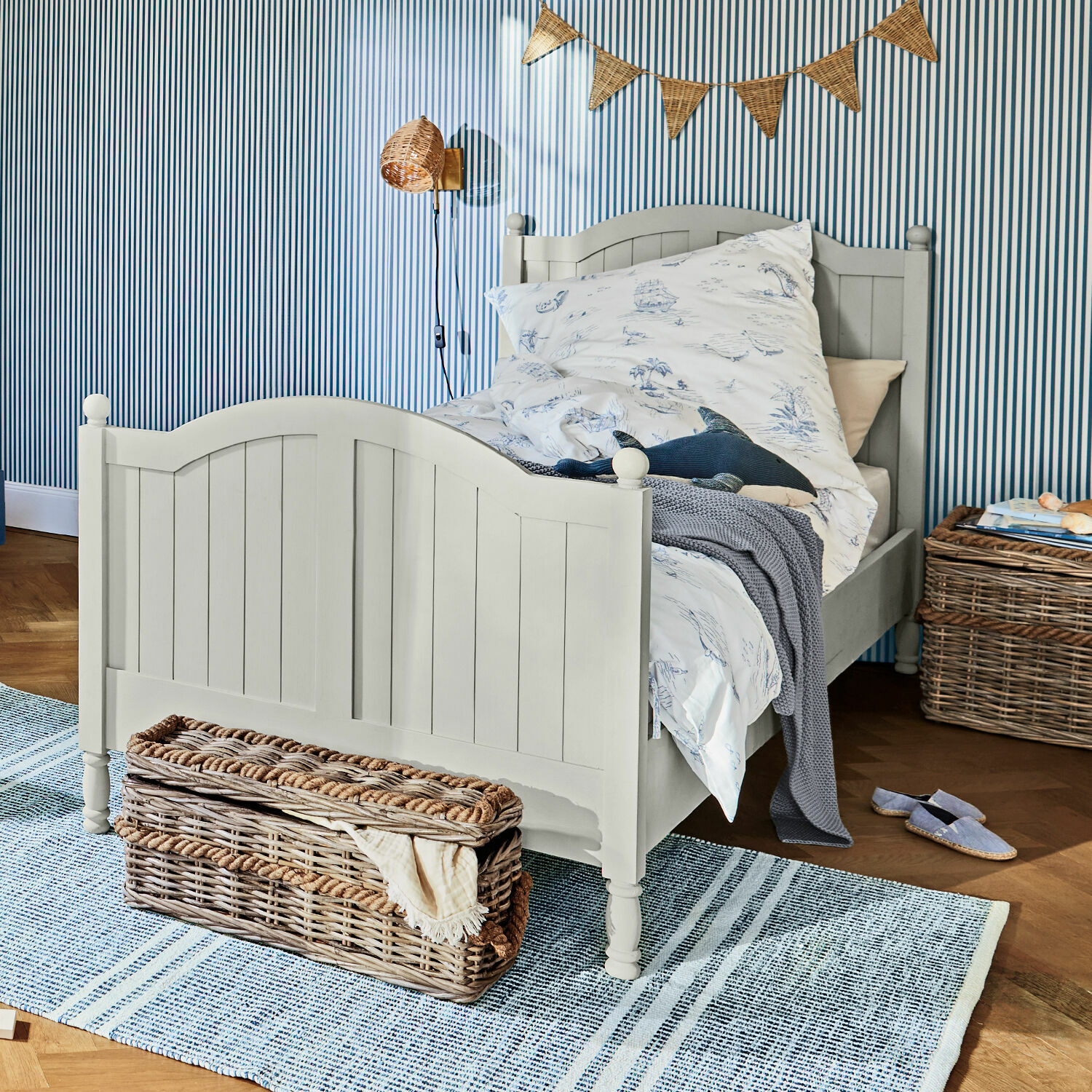 Cama Alarion Cama infantil de madera gris con ropa de cama de motivos marinos y cesta de mimbre sobre alfombra