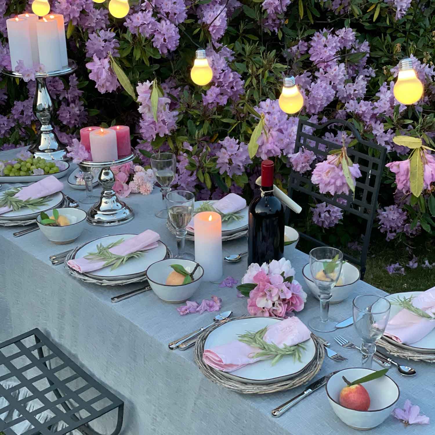 Mesa de jardín elegantemente puesta con servilletas rosas, velas y botella de vino frente a flores moradas