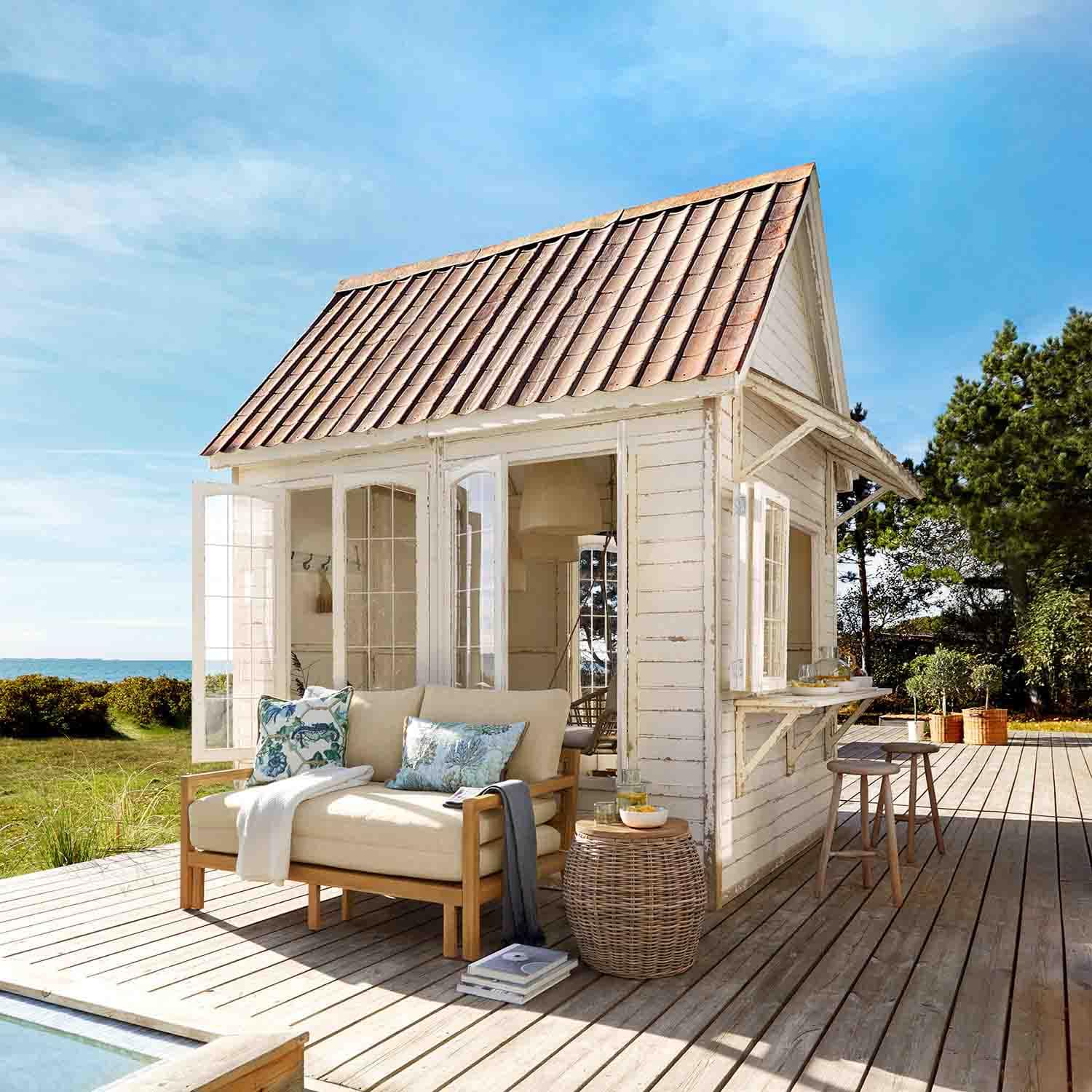 Caseta de madera blanca con techo rojo en terraza de madera con sofá beige y mesa de mimbre