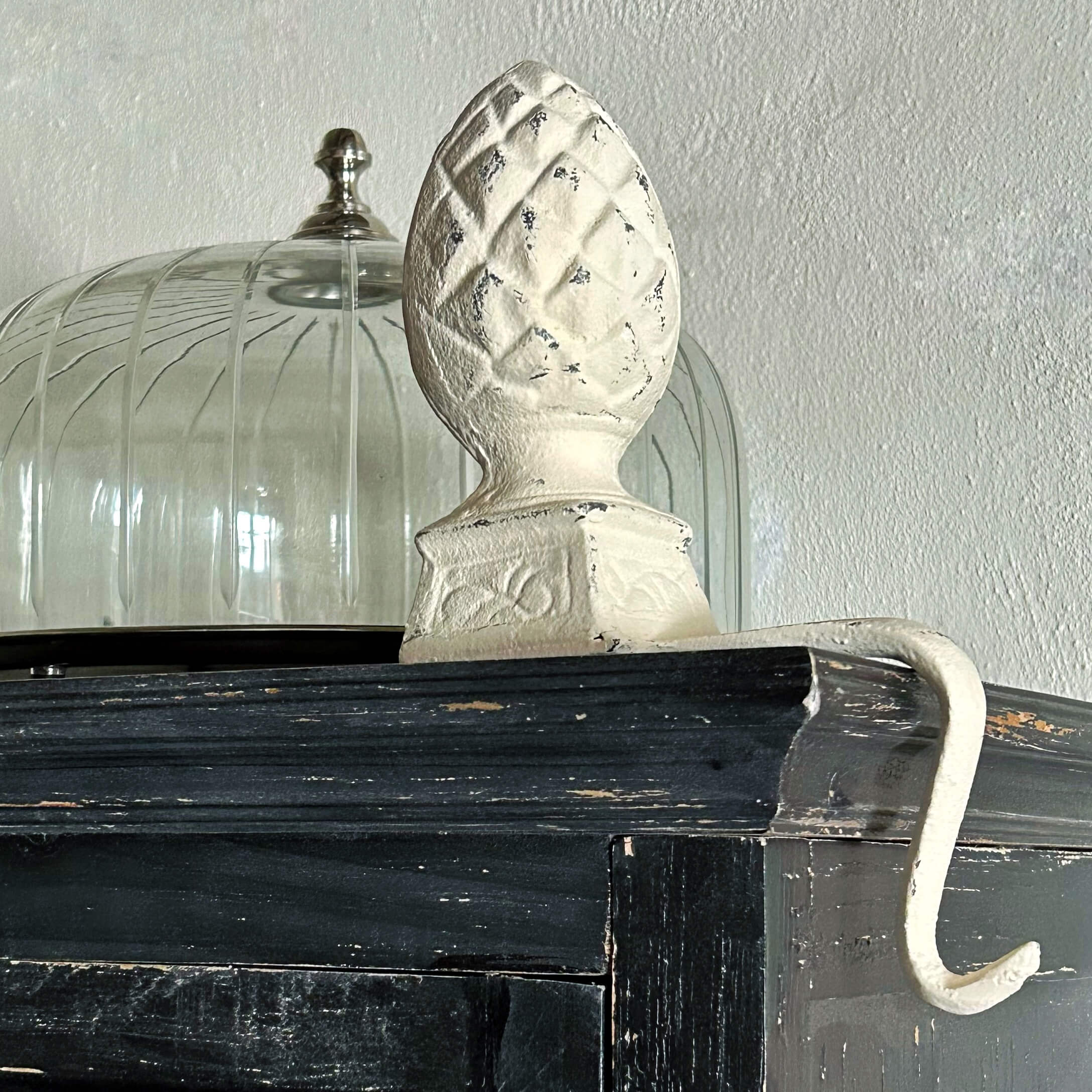 Gancho metálico blanco con remate decorativo en forma de piña sobre estante de madera negra