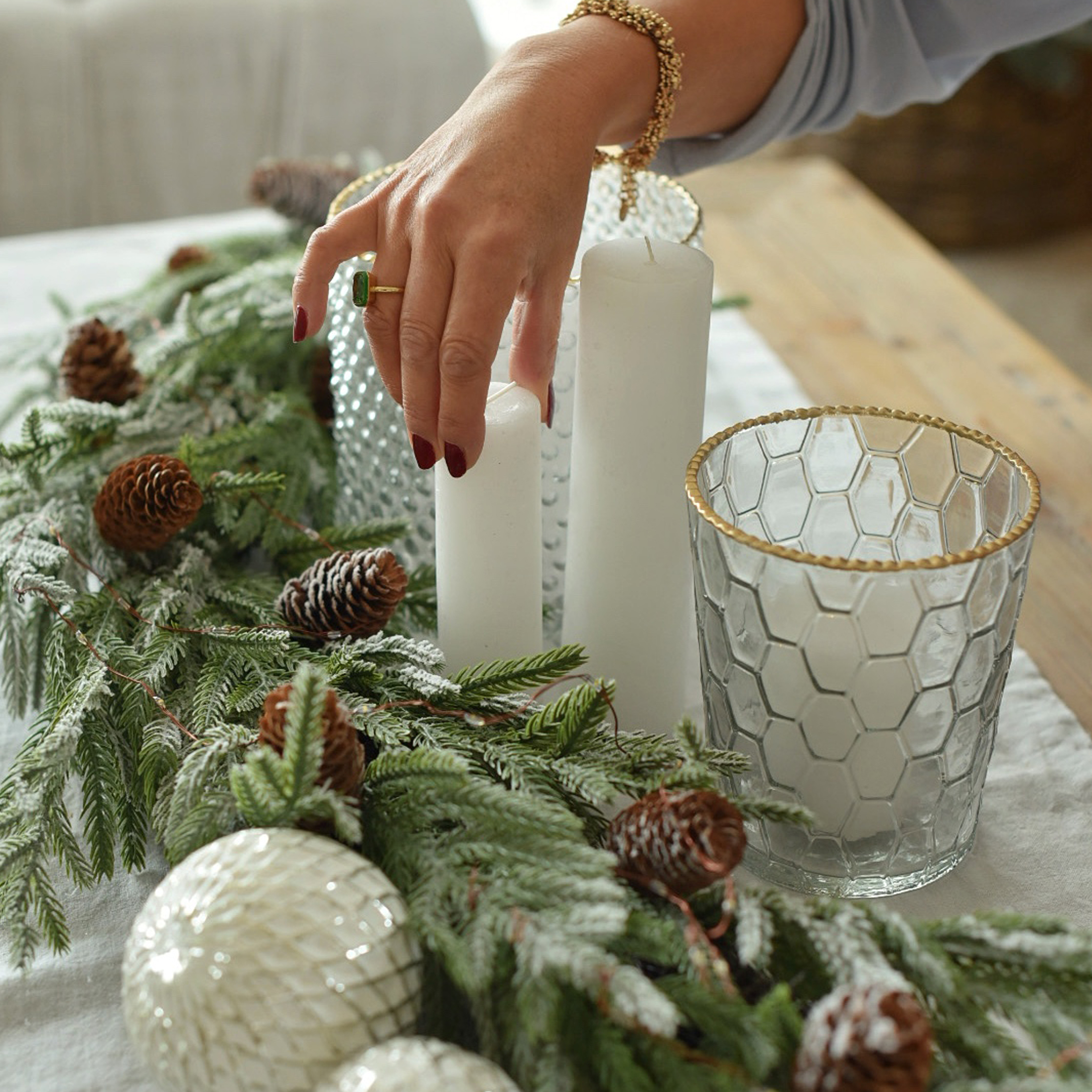 Decoración navideña con velas blancas, portavelas de cristal con borde dorado y ramas verdes con piñas