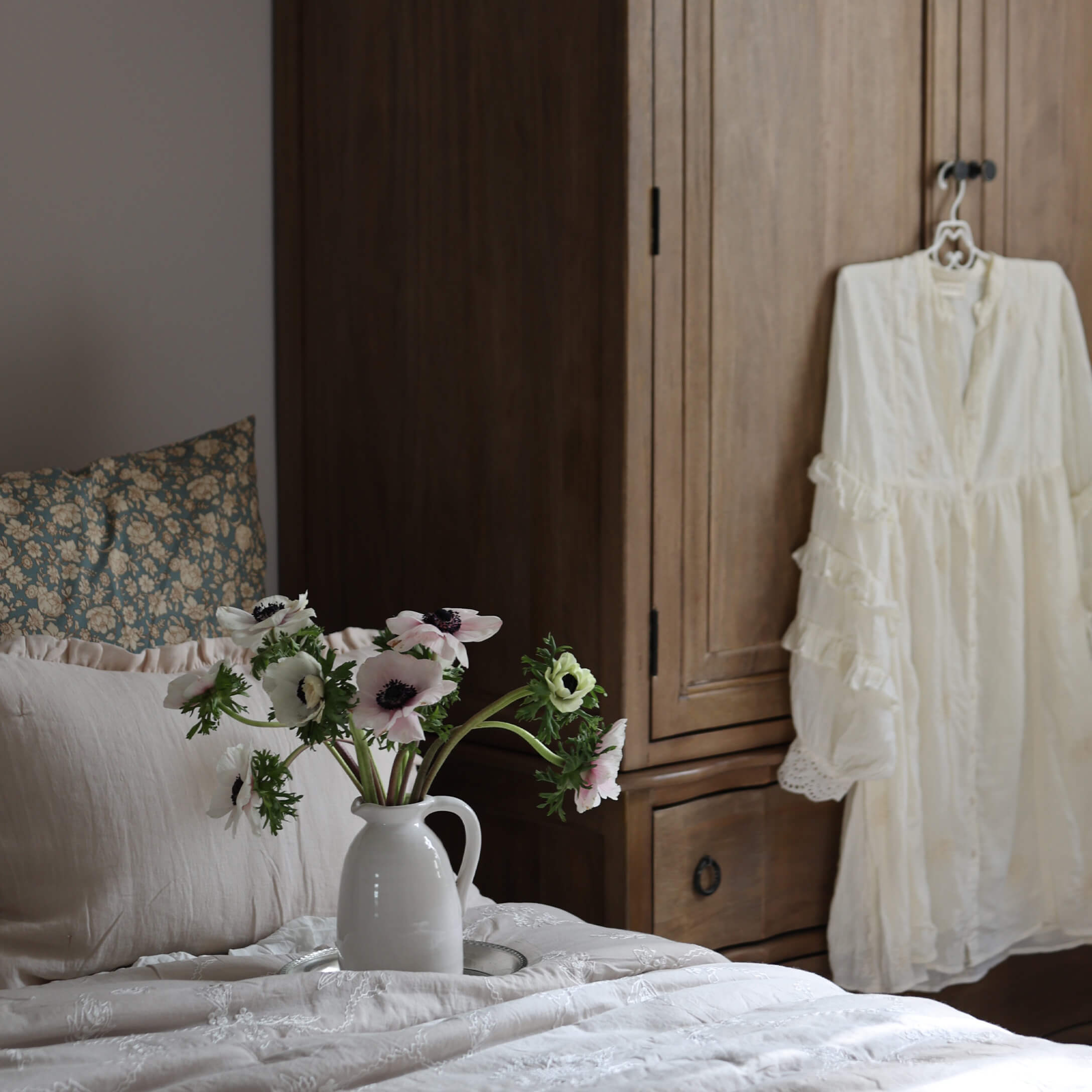 Jarra de cerámica blanca con flores sobre cama con ropa clara y armario de madera al fondo
