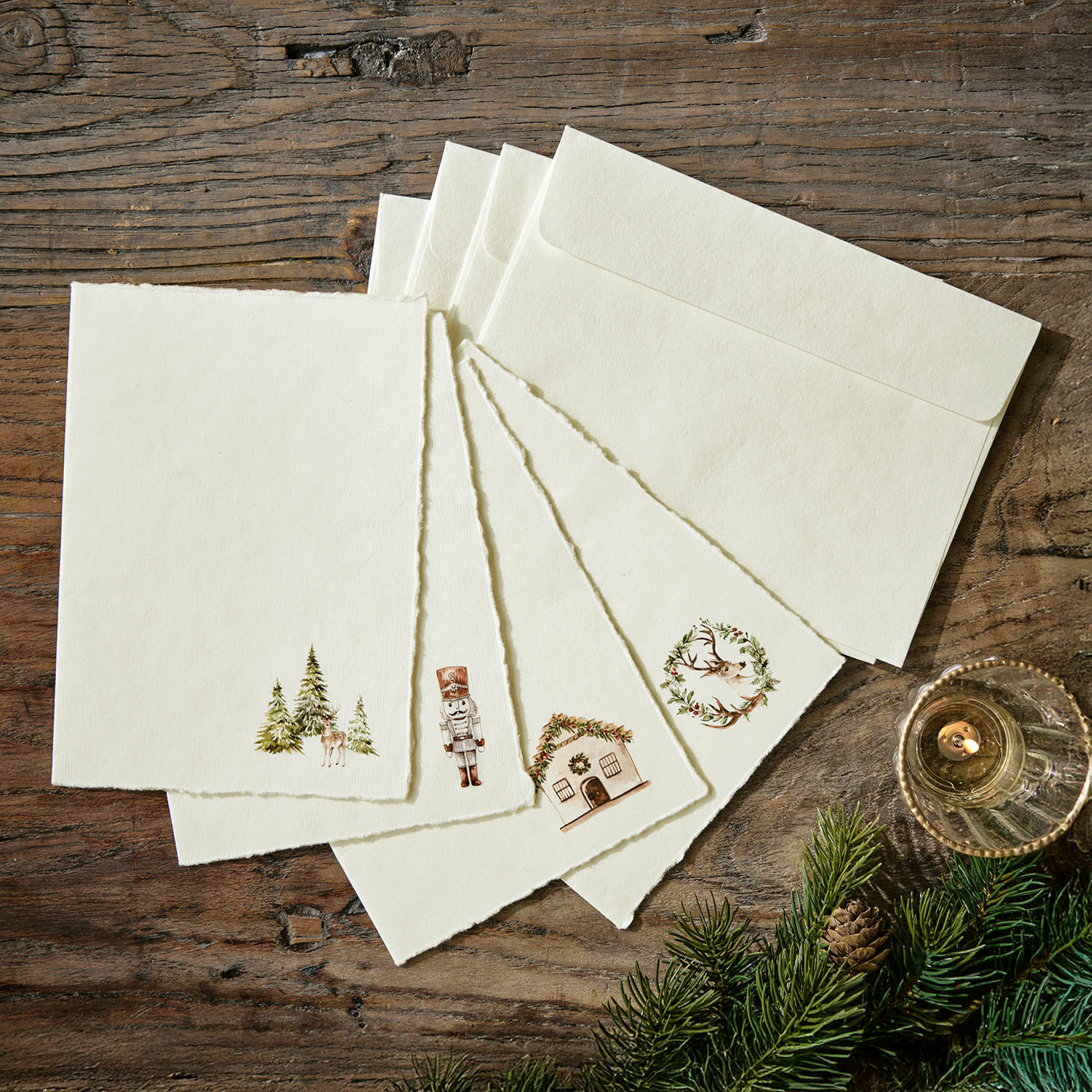 Set de 4 tarjetas de felicitación Lorandis Conjunto de tarjetas y sobres crema con motivos navideños sobre mesa de madera con ramas de abeto