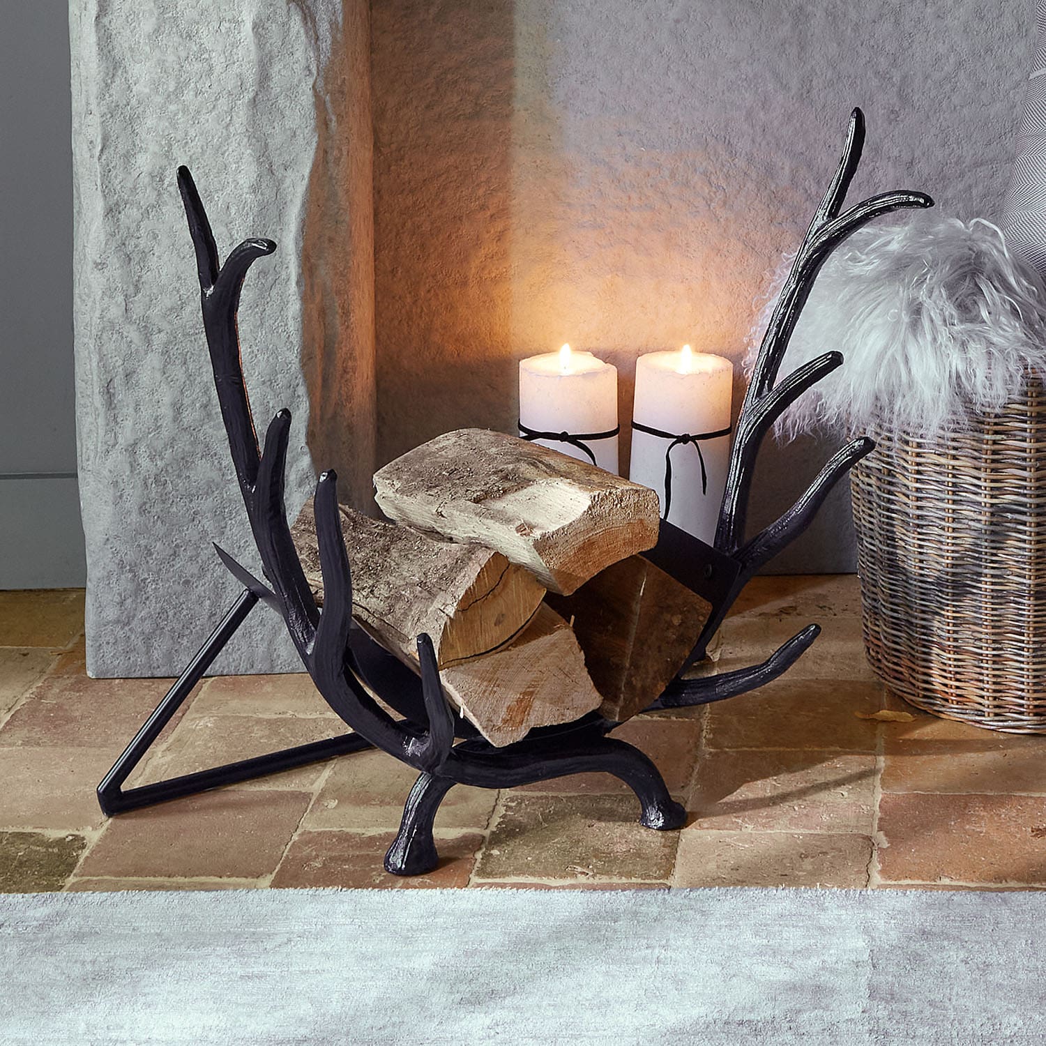 Soporte para leña de metal negro con forma de cuernos, con troncos y velas encendidas al fondo