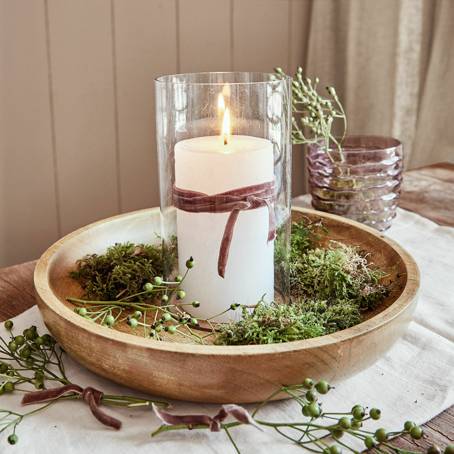 Vela blanca en vaso de cristal con lazo de terciopelo sobre bandeja de madera con musgo verde