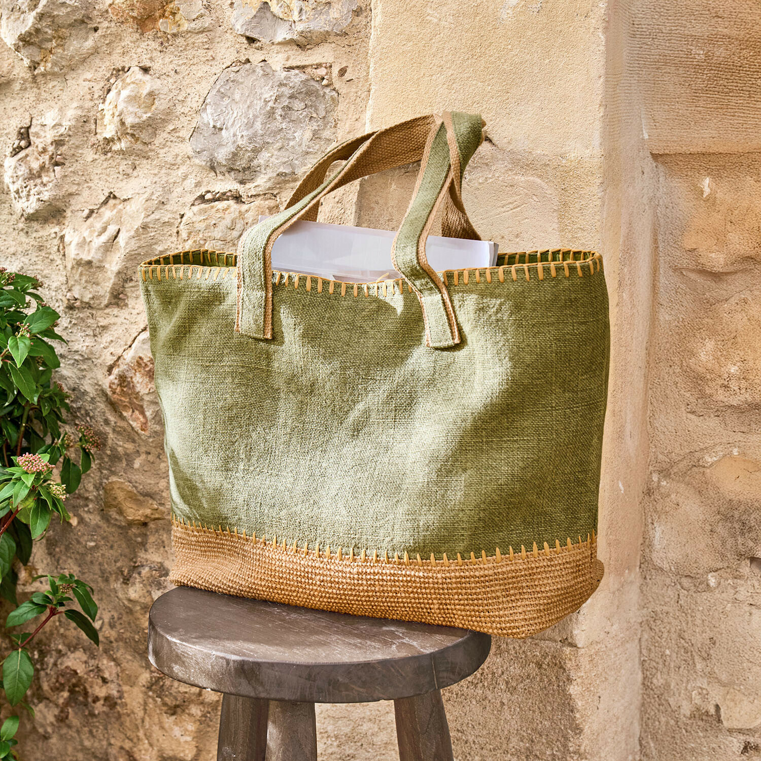 Bolso Yarrowton Bolsa verde de yute con acabado marrón sobre un taburete de madera junto a una pared de piedra