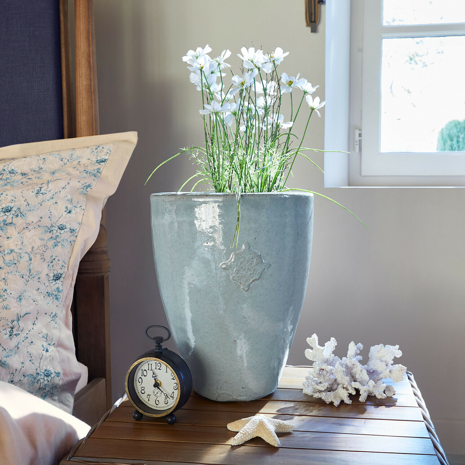 Macetero Serenzia Maceta de cerámica gris con flores blancas sobre mesa de madera junto a reloj despertador y decoraciones marinas