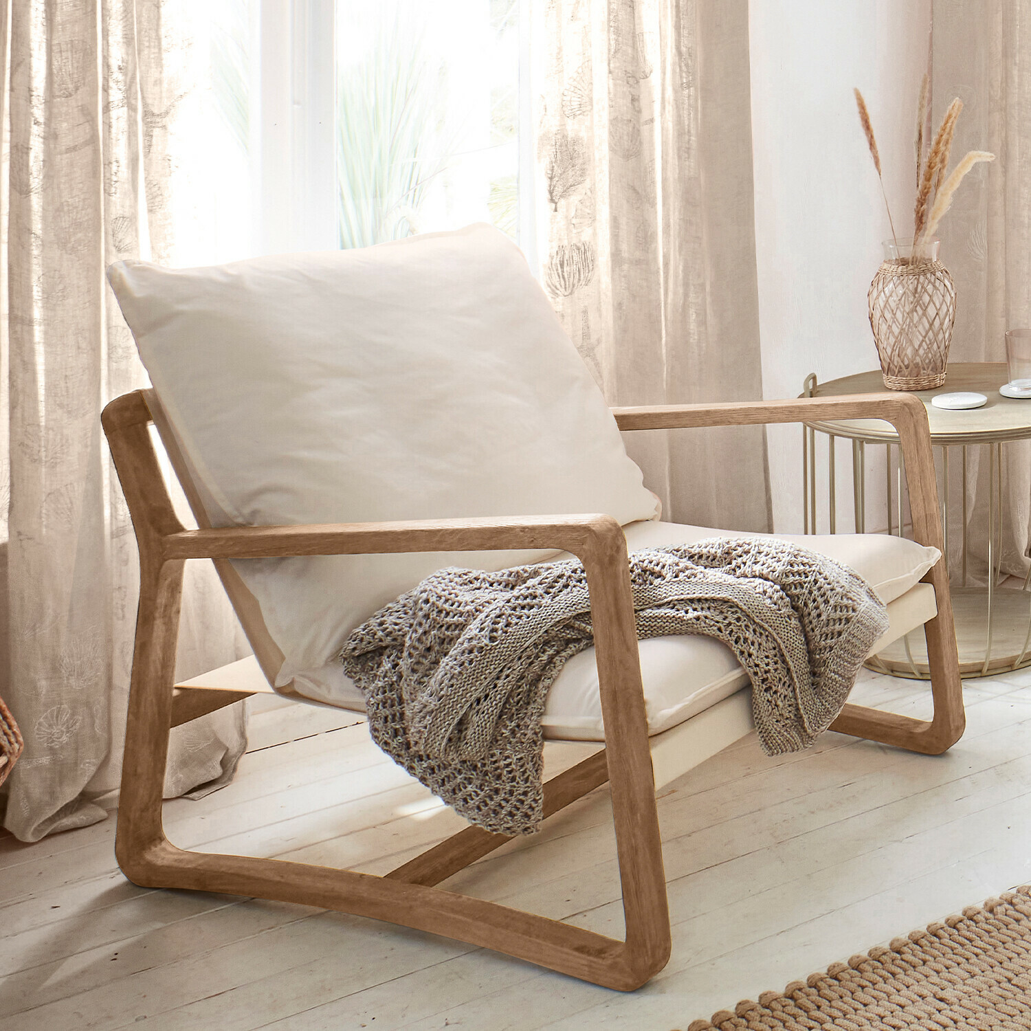 Sillón Jowna Sillón de madera clara con cojín crema y manta tejida gris en sala luminosa