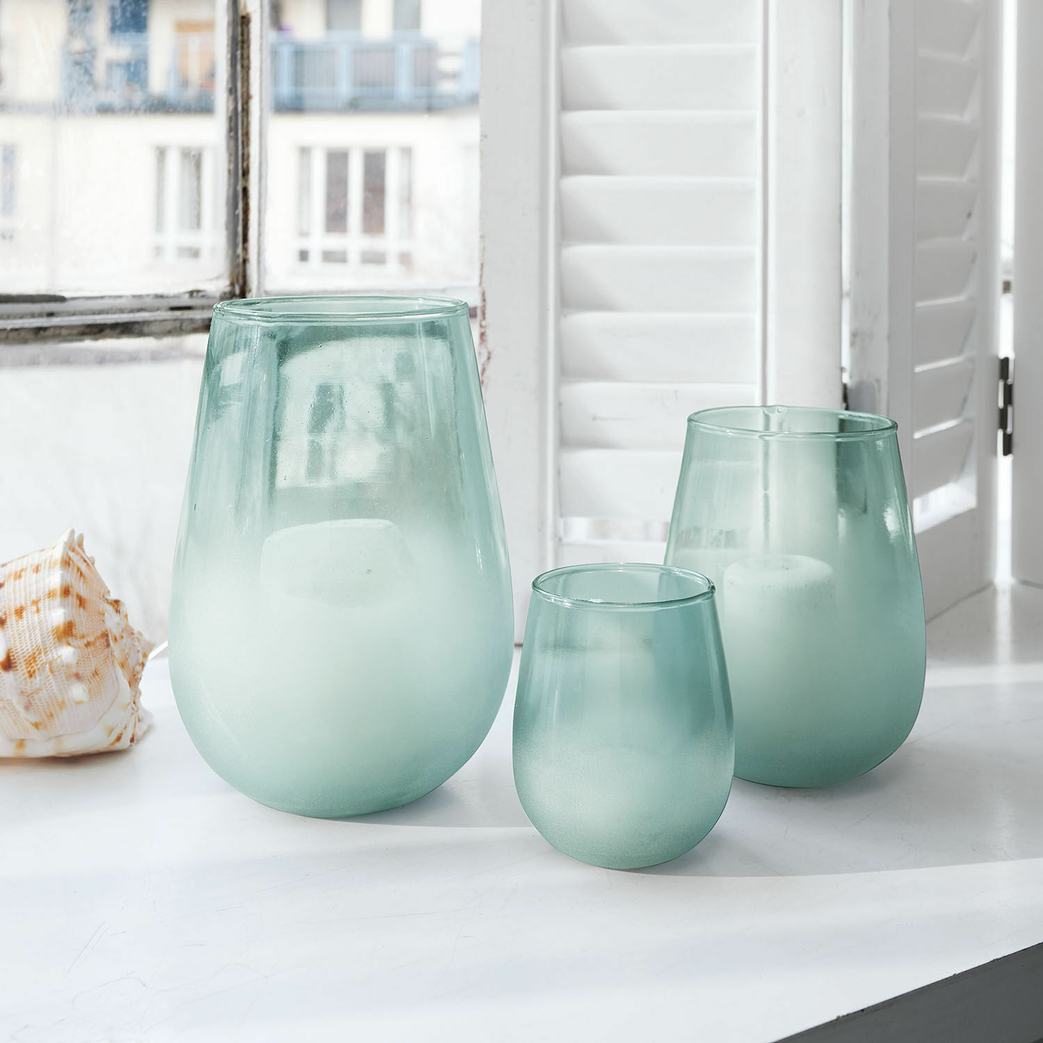 Conjunto de tres portavelas de vidrio mate verde sobre un alféizar blanco con velas blancas