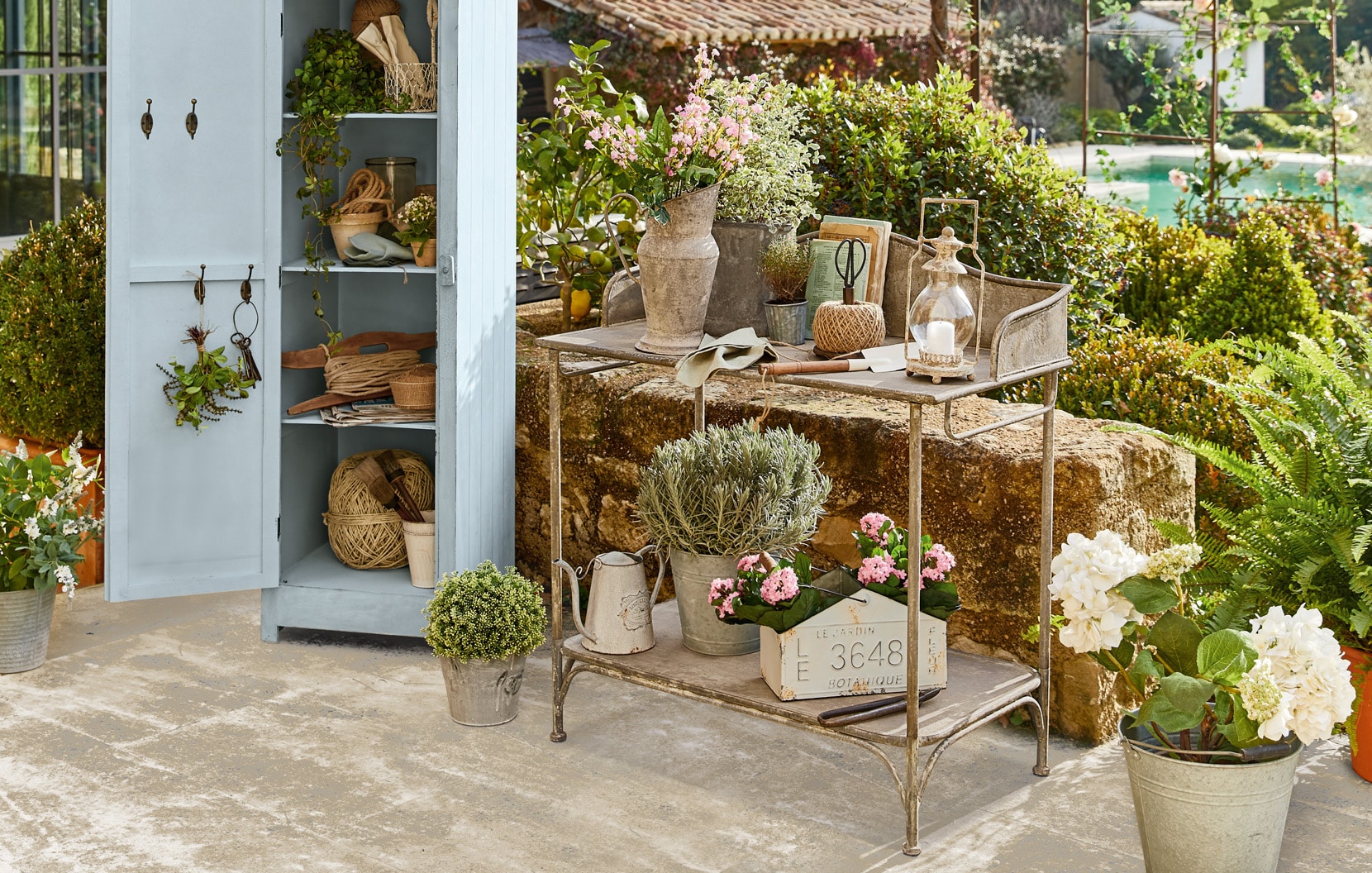Carrito de jardín de metal con decoraciones shabby chic y flores en patio con armario azul.