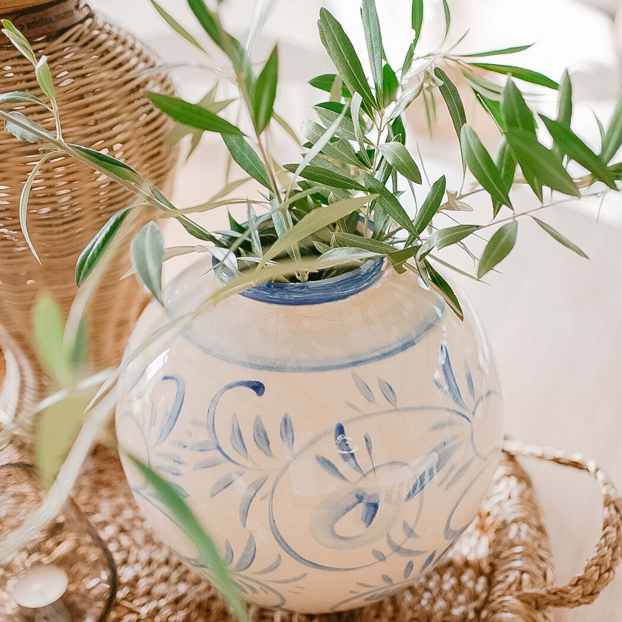 Jarrón de cerámica blanca con diseño azul y hojas verdes sobre bandeja de mimbre, decoración primaveral