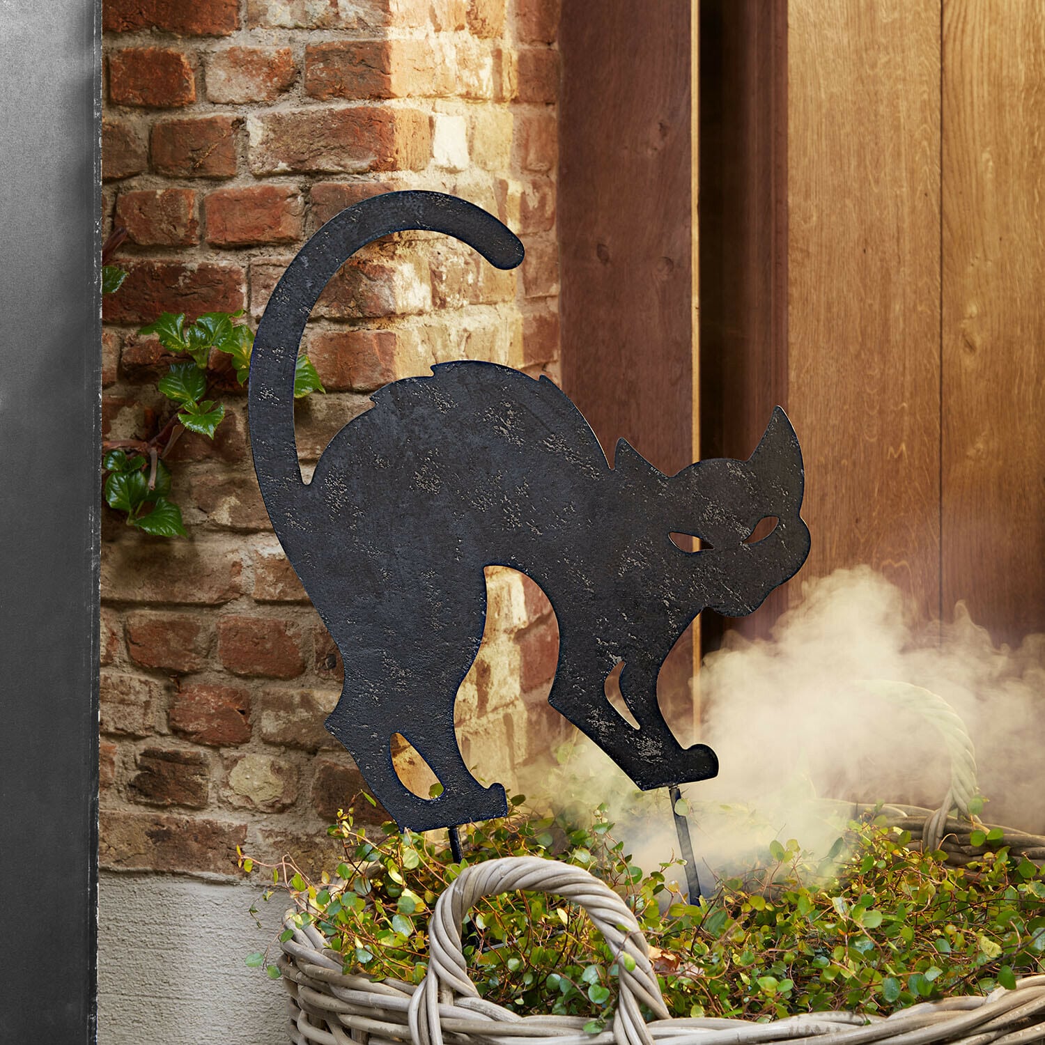 Estaca de jardín Rénelise Silueta decorativa de gato negro de metal en cesta con plantas verdes frente a pared de ladrillo