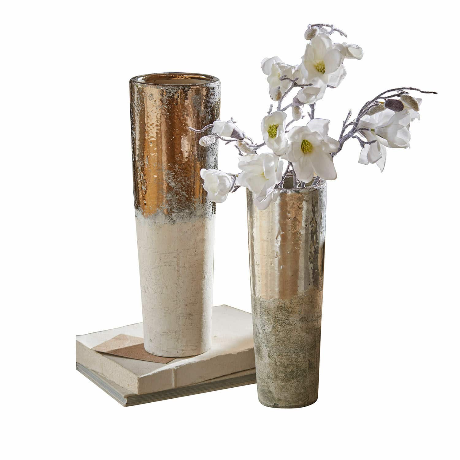 Set de 2 jarrones Tameena Dos jarrones metálicos altos en tonos dorado y plateado sobre libros con flores blancas decorativas