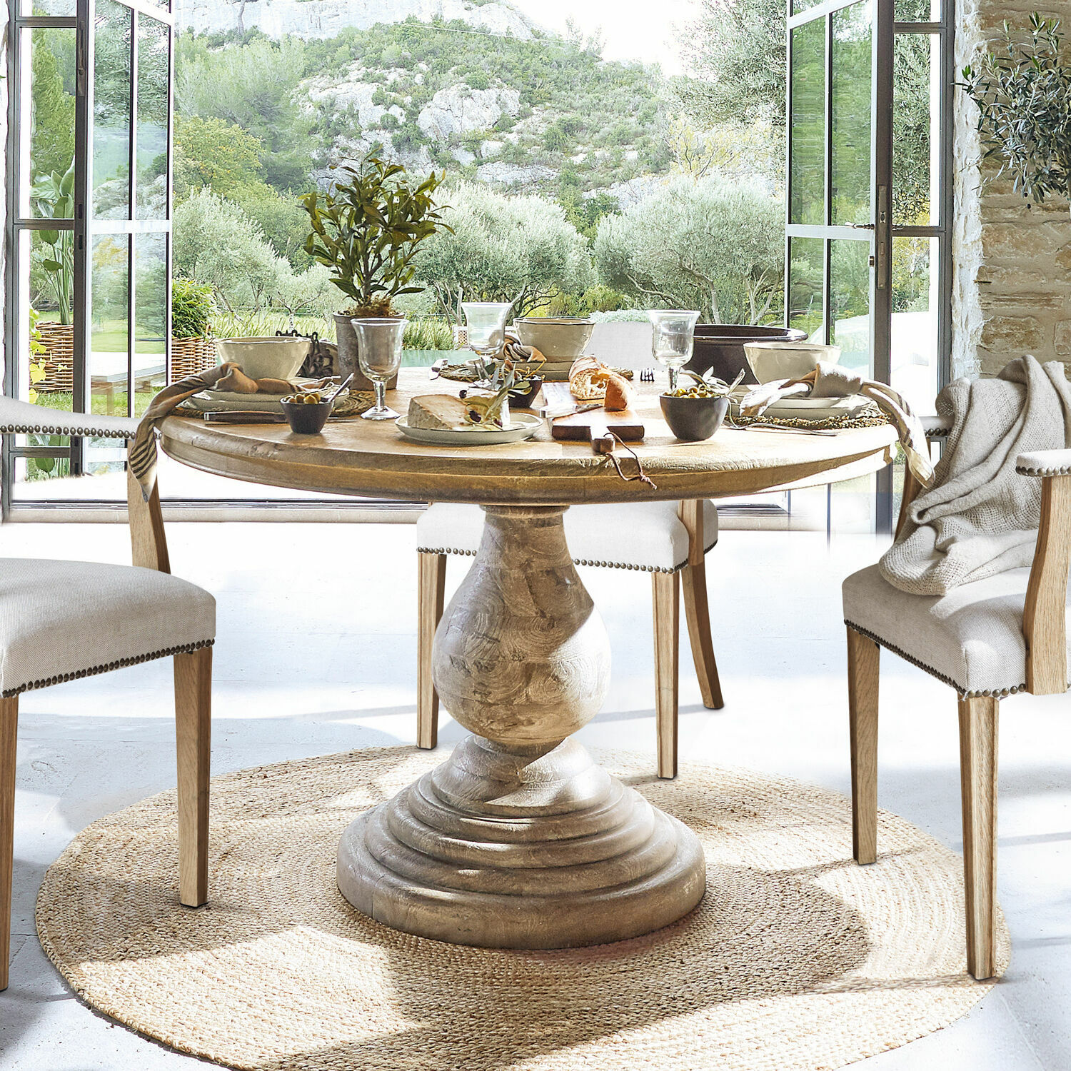 Mesa Orlin Mesa redonda de madera con base decorativa sobre alfombra natural en comedor luminoso con vista al jardín