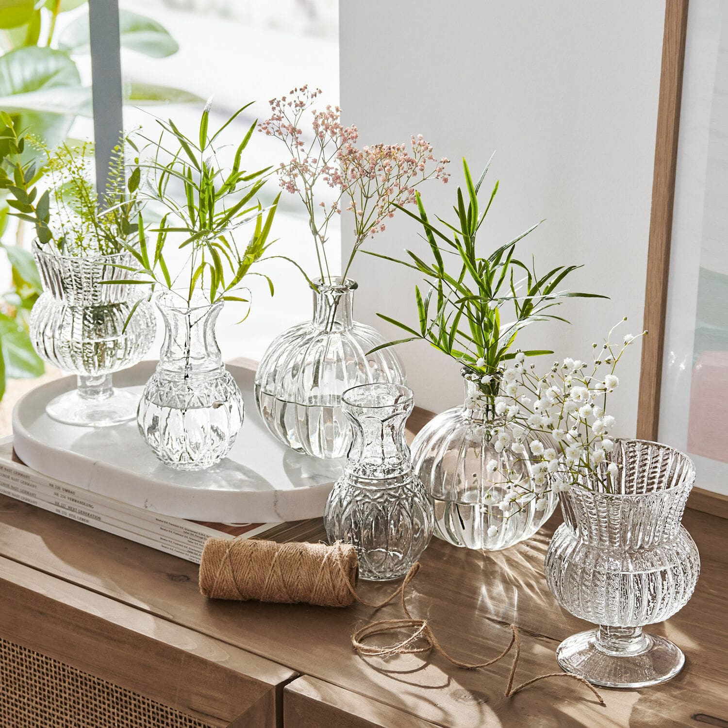 Set de 6 jarrones Amber Conjunto de jarrones de vidrio transparente con flores verdes y blancas sobre mueble de madera