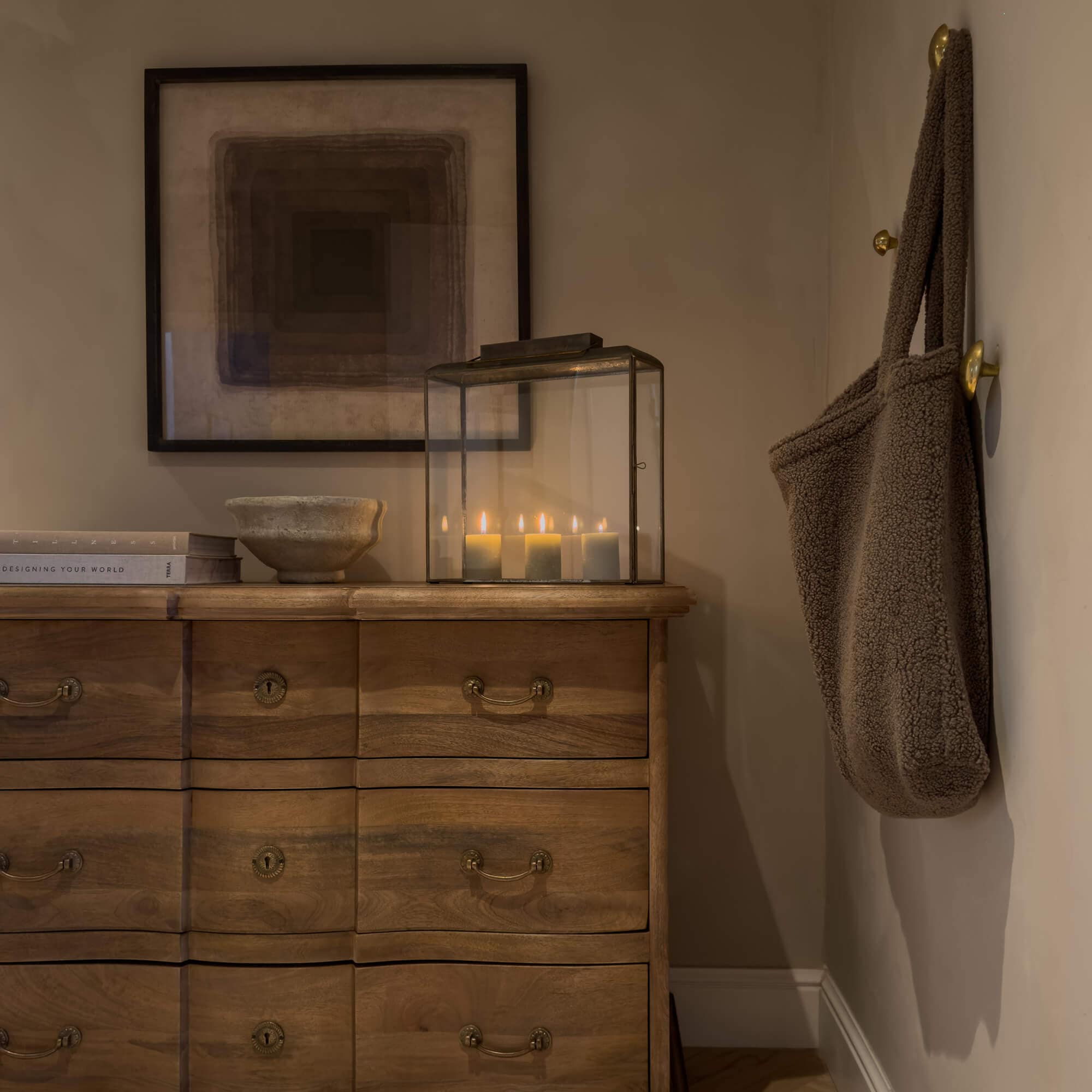 Cómoda de madera con tiradores de latón, farol de cristal con velas encendidas y bolso marrón de material suave colgado en la pared
