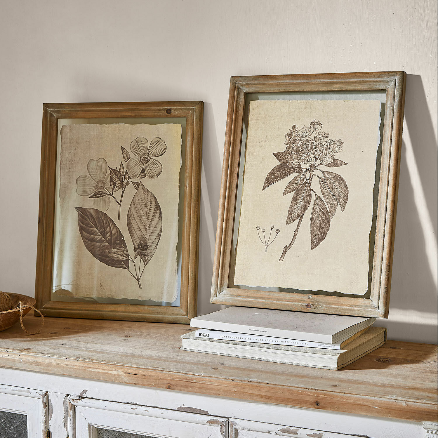 Set de 2 cuadros Guyra Dos marcos de madera rústica con dibujos botánicos de flores sobre fondo beige sobre una mesa de madera