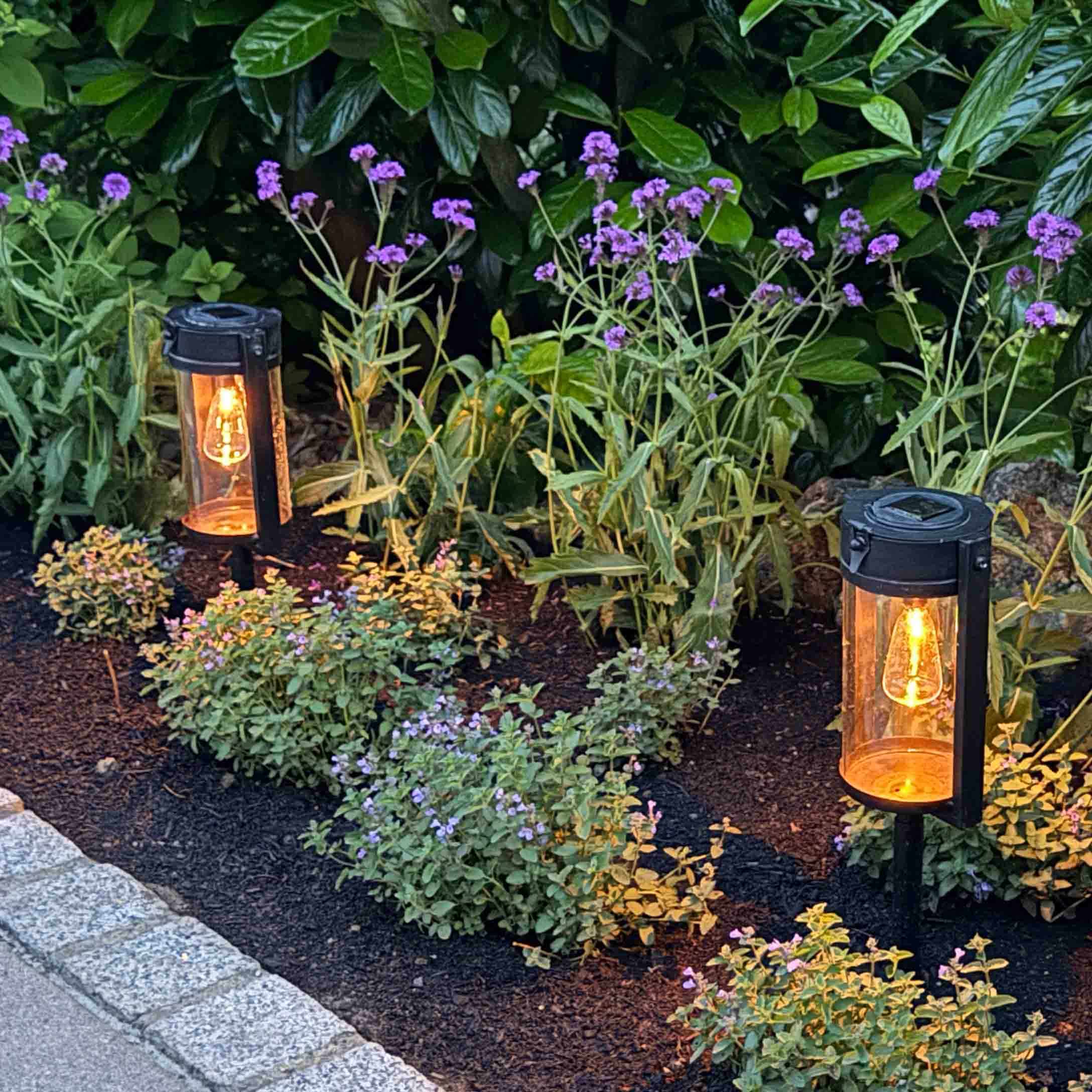 Iluminación de jardín de metal negro con luz cálida entre plantas verdes y flores moradas
