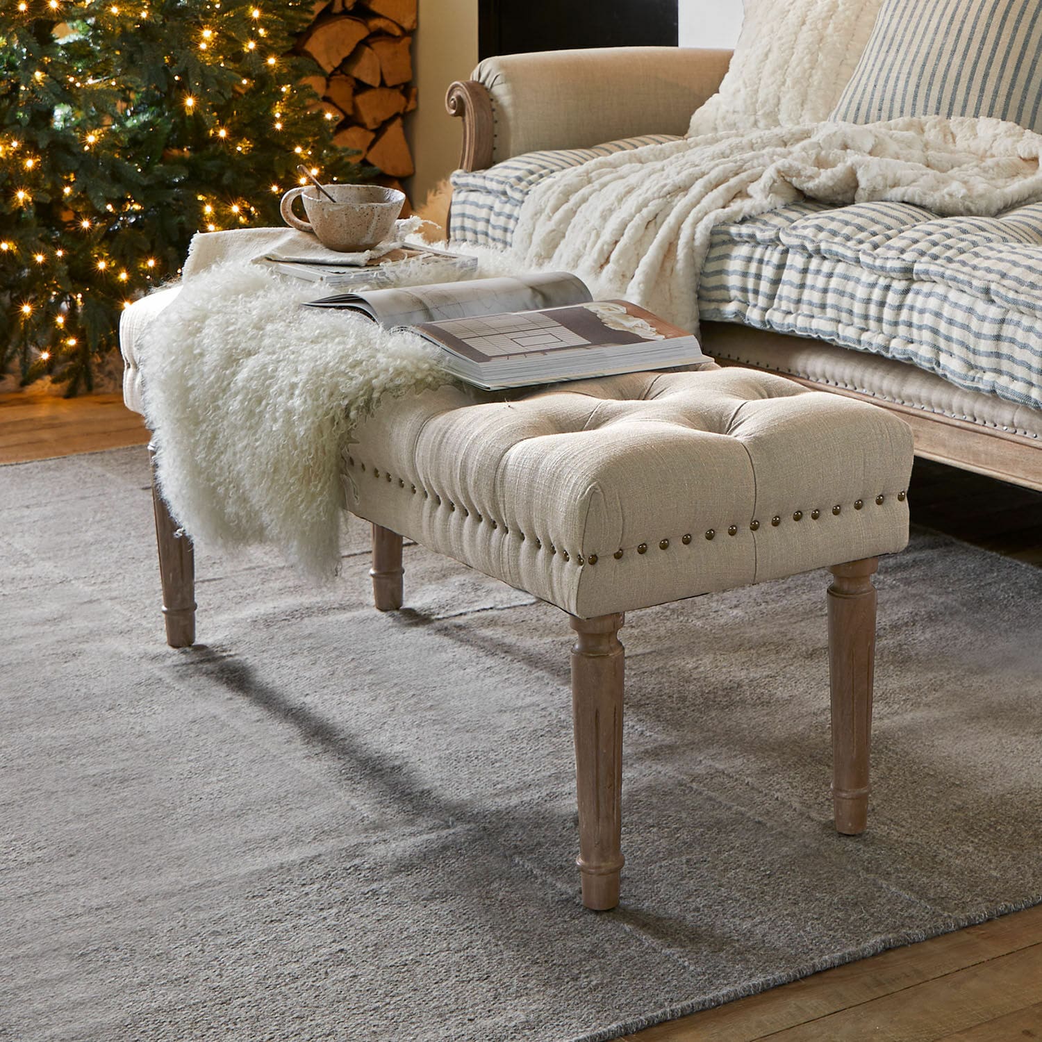 Banco beige acolchado con patas de madera sobre alfombra gris en sala acogedora con árbol de Navidad