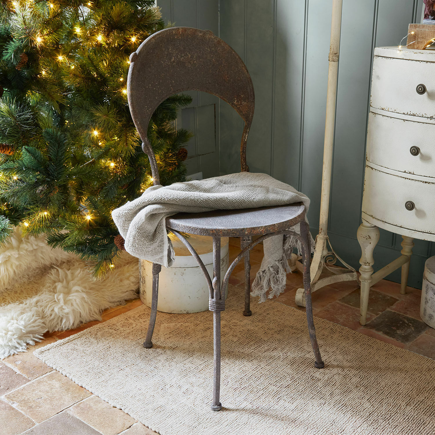 Silla Delivora Silla rústica de metal oxidado con manta gris junto a árbol de Navidad iluminado y alfombra