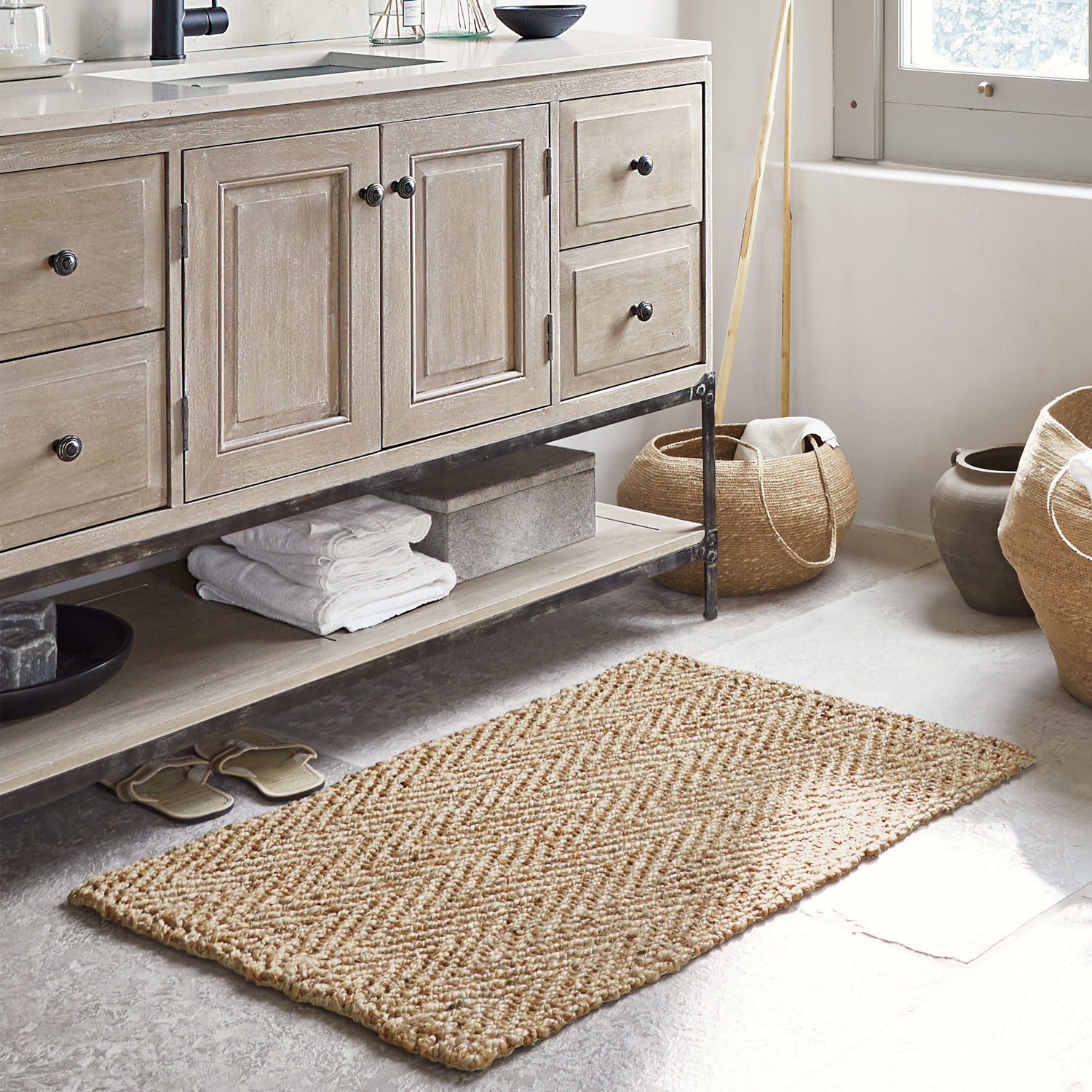 Alfombrilla de baño Daoukro Alfombra beige de yute con patrón espiga en baño con mueble de madera y cestas
