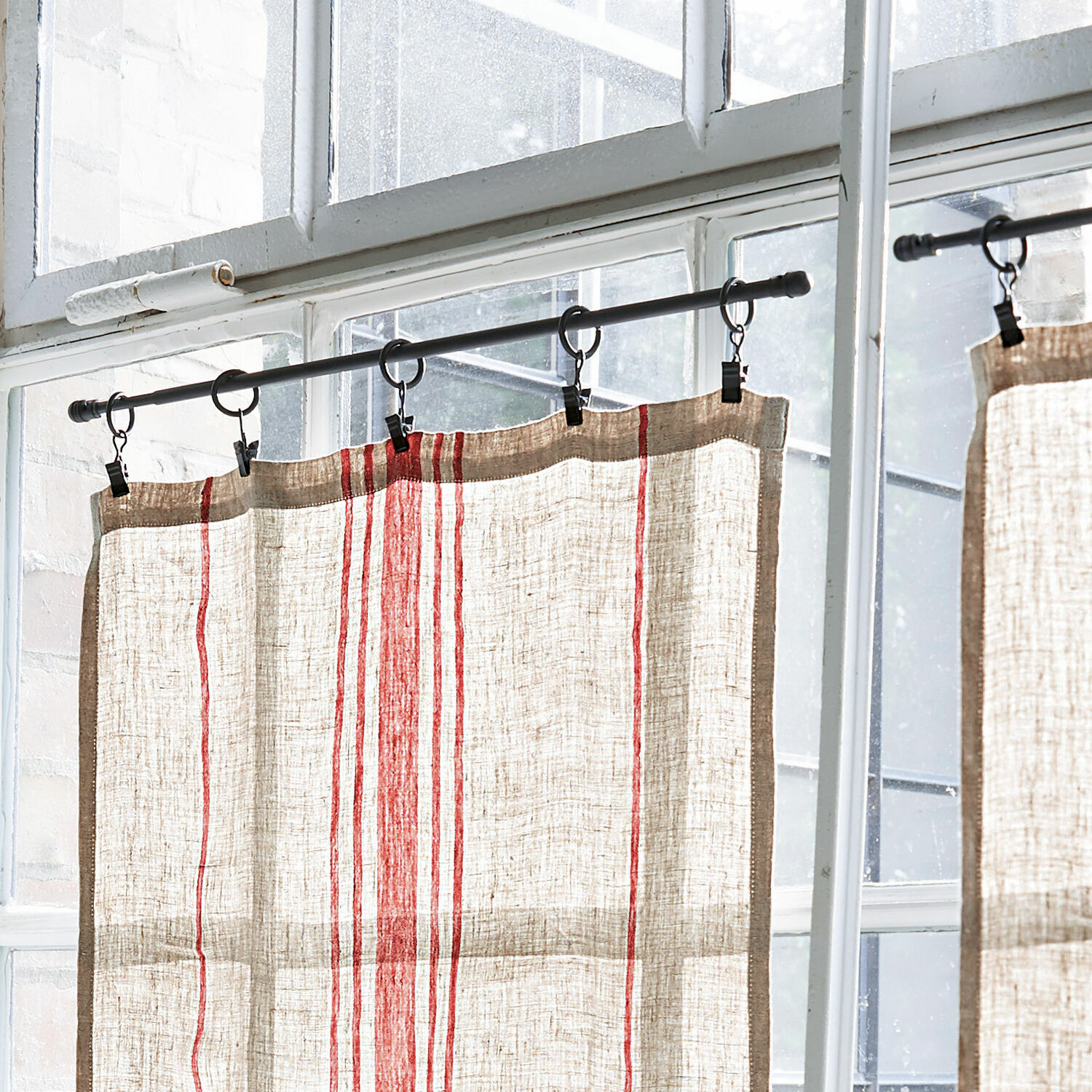Cortina de lino color natural con rayas rojas colgada en barra metálica frente a ventana