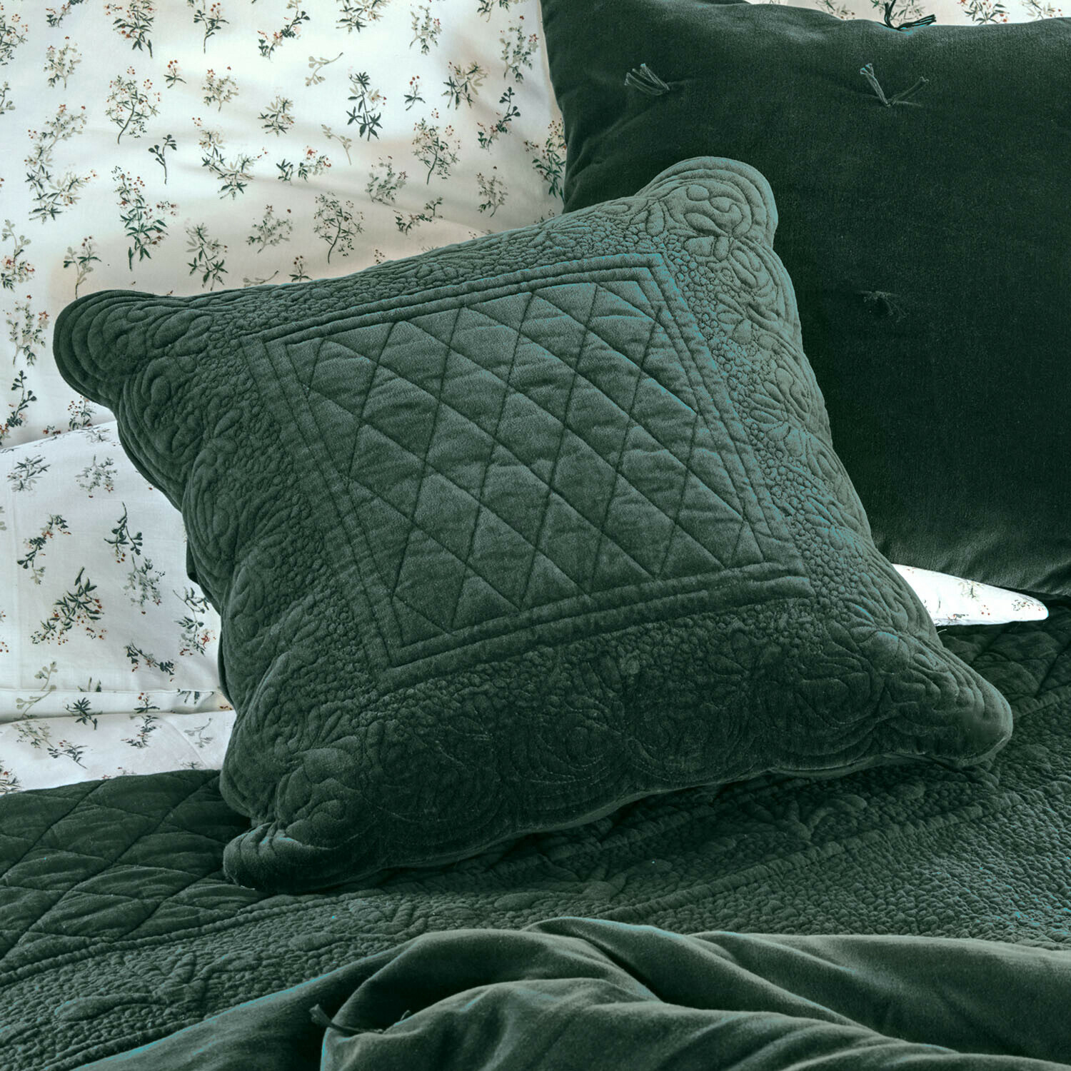 Cojín Lunévois Cojín verde de terciopelo con diseño acolchado sobre cama con ropa de cama floral y manta