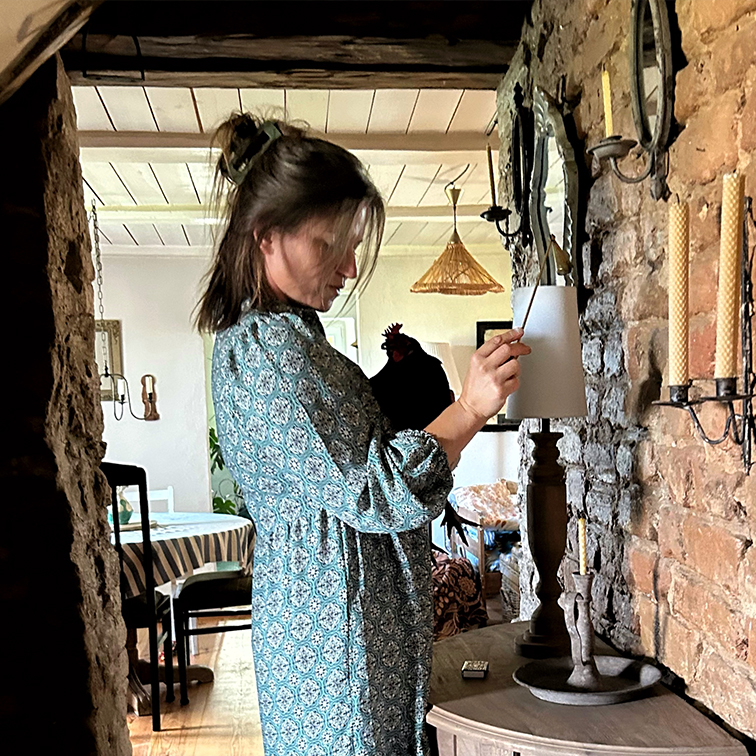 Mujer con vestido azul estampado encendiendo lámpara en interior rústico con pared de ladrillo y velas