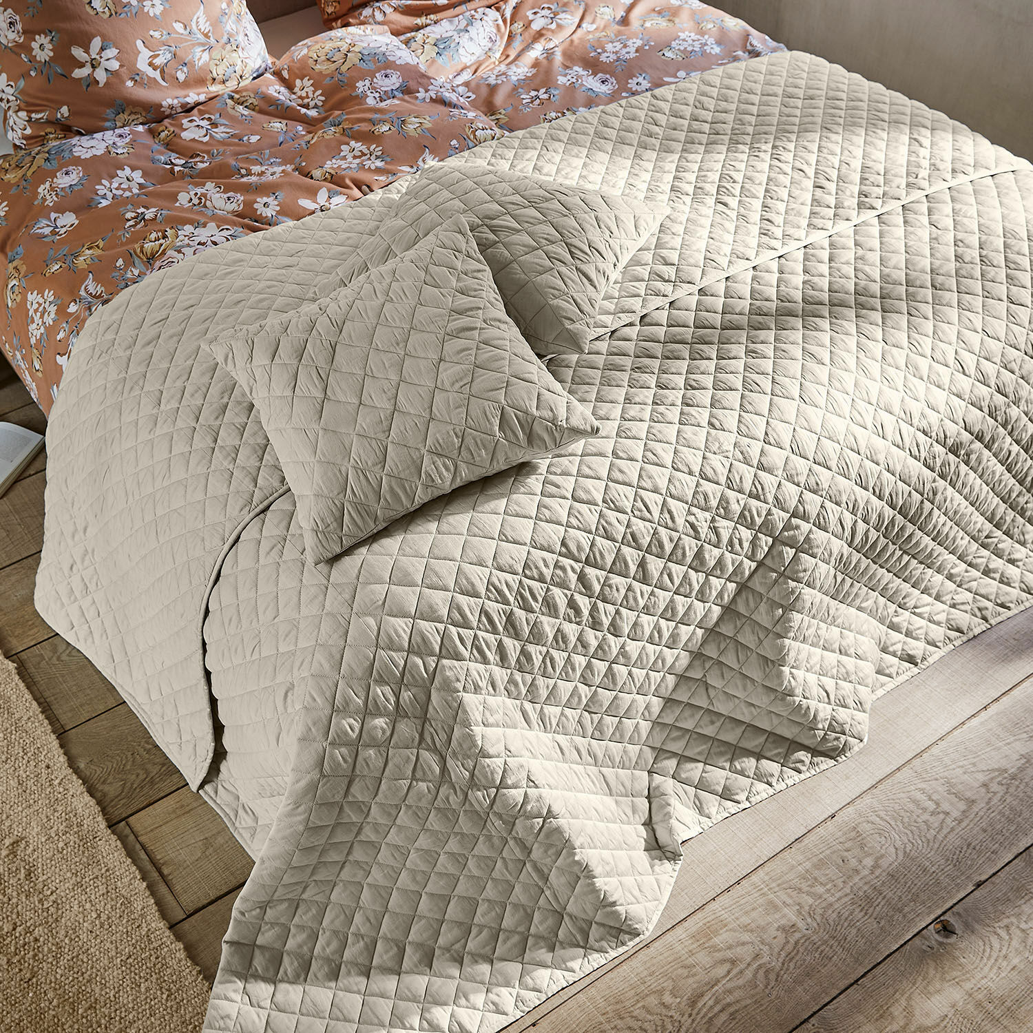 Colcha Jolique Colcha beige acolchada con cojines sobre cama de madera y ropa de cama floral