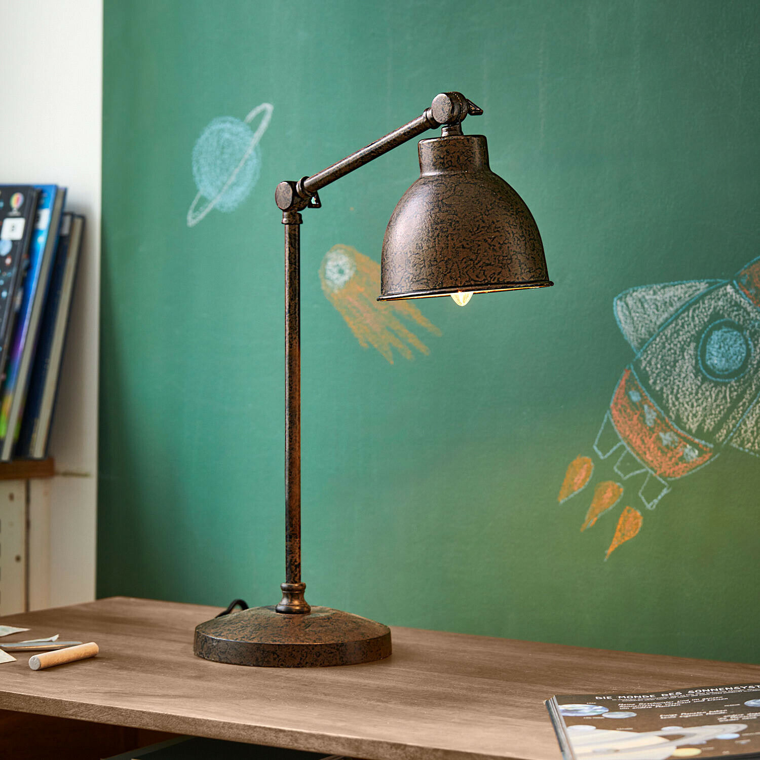 Lámpara de mesa de metal con acabado oxidado sobre escritorio de madera frente a pizarra verde con dibujos espaciales