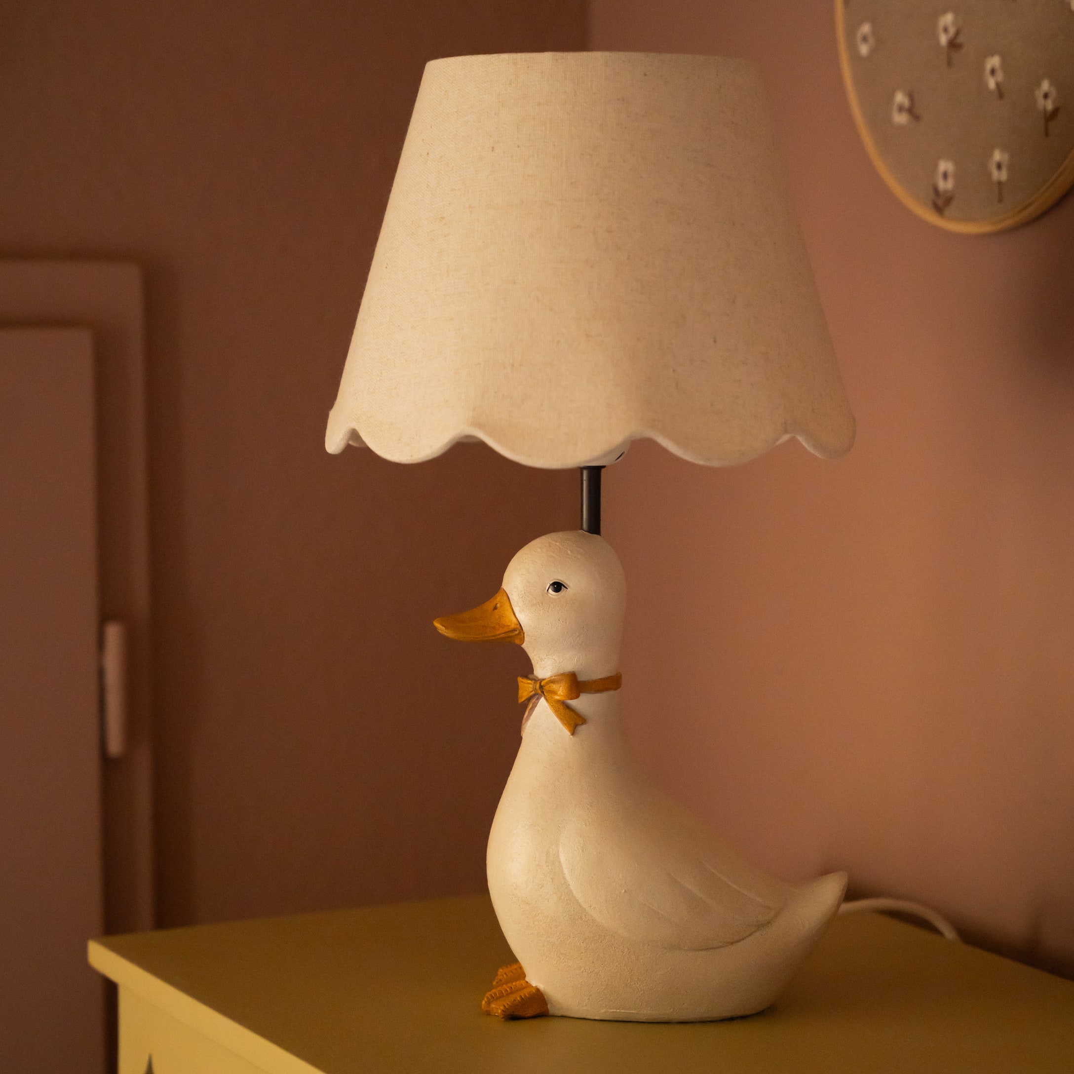 Lámpara de mesa con base en forma de pato blanco, pantalla beige y lazo naranja decorativo