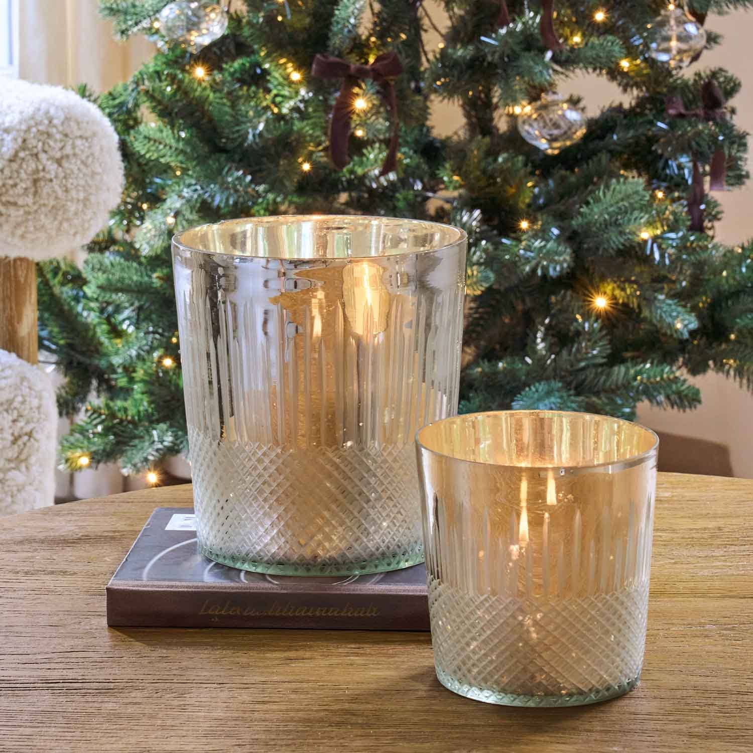 Dos portavelas de vidrio con acabado metálico sobre mesa de madera junto a un árbol de Navidad decorado