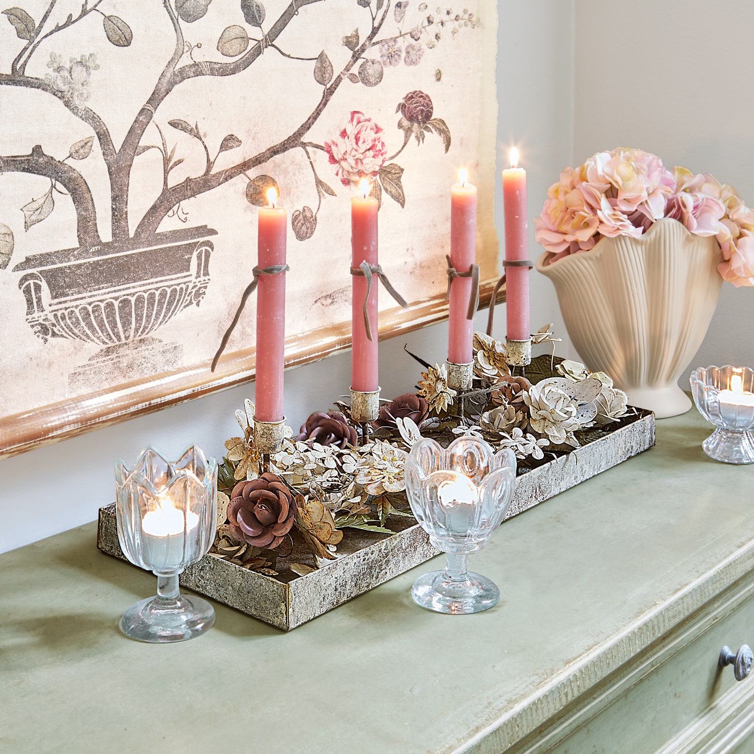 Candelabro decorativo con flores metálicas y velas rosas sobre cómoda verde con jarrón