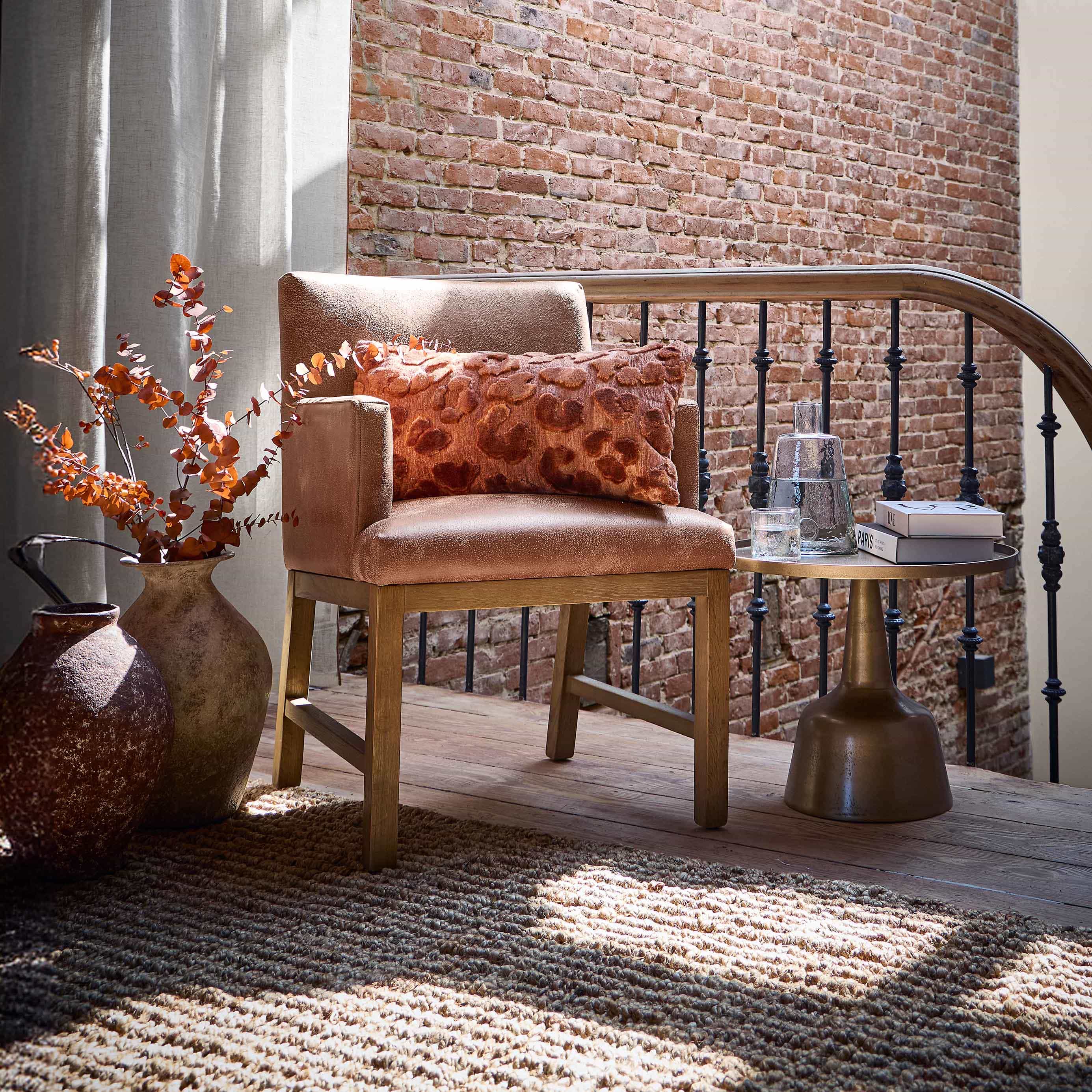 Sillón beige con base de madera y cojín naranja frente a pared de ladrillo y cortinas