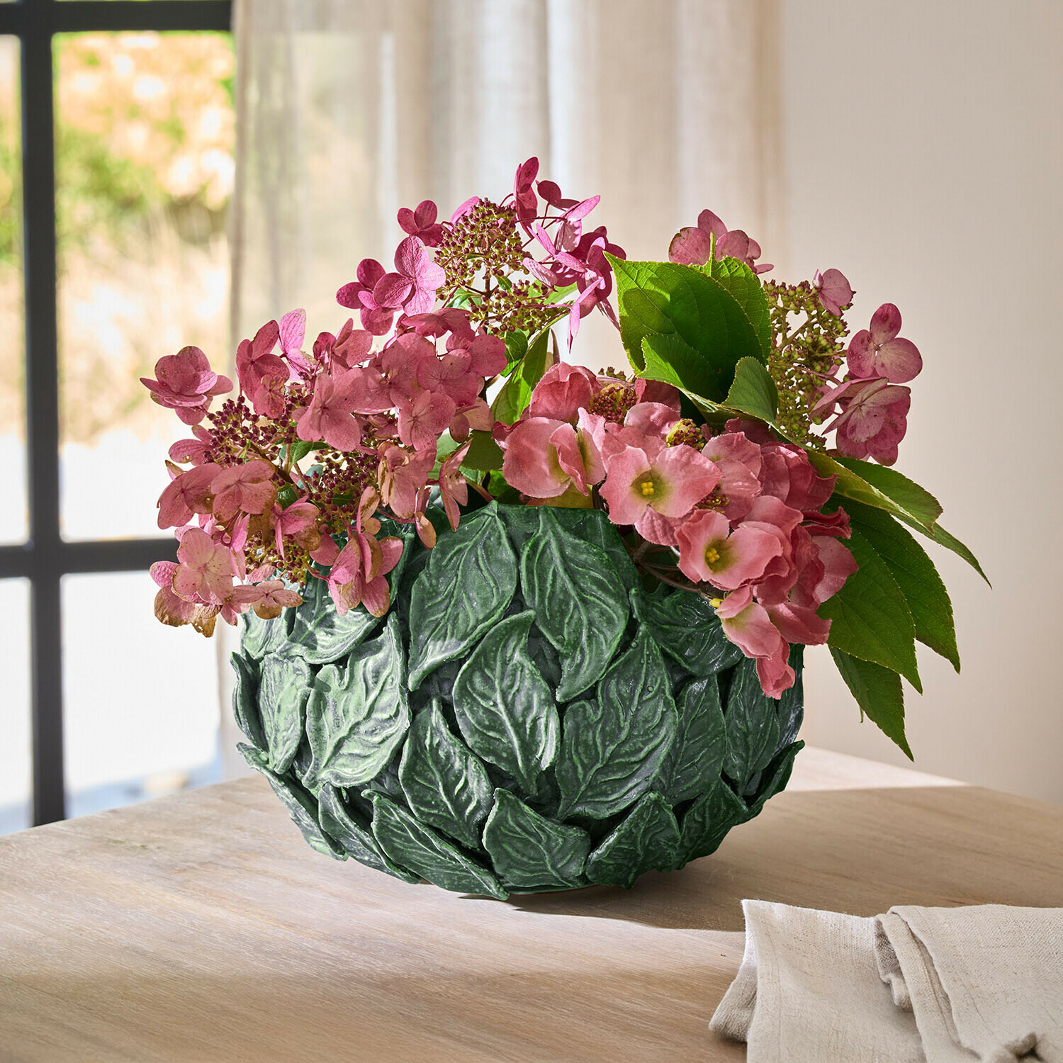 Jarrón decorativo verde con hojas y flores rosas sobre mesa de madera en interior luminoso
