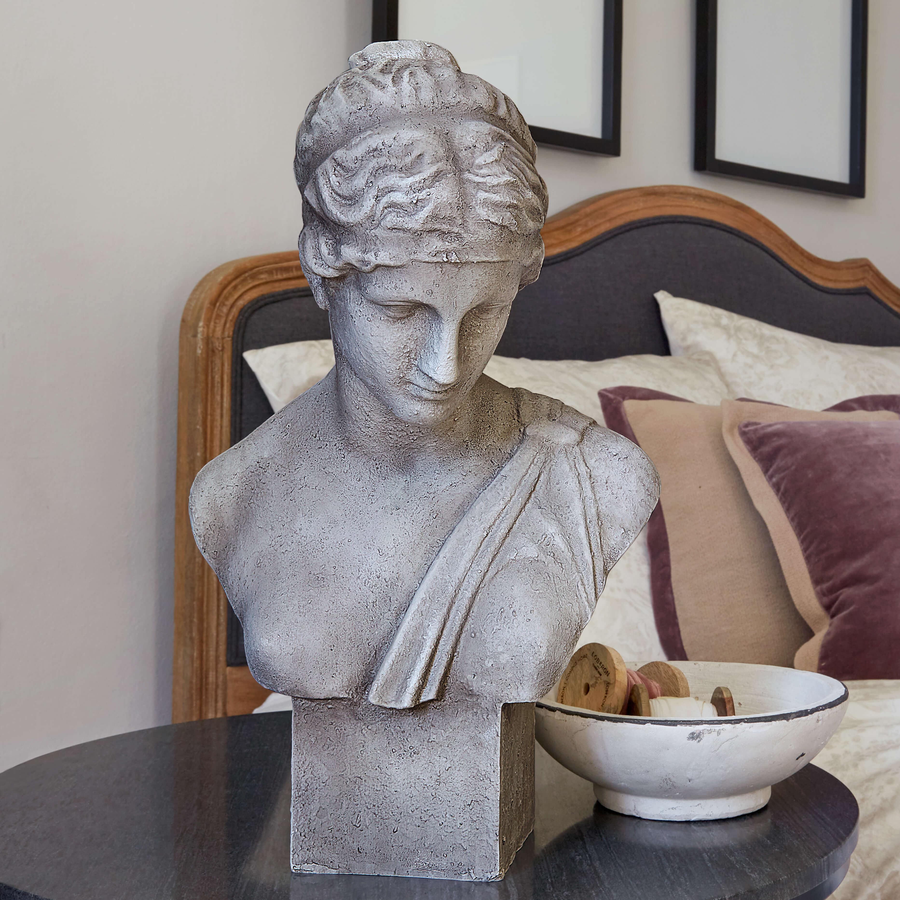 Busto de piedra gris sobre mesa junto a un cuenco blanco con carretes de madera en dormitorio