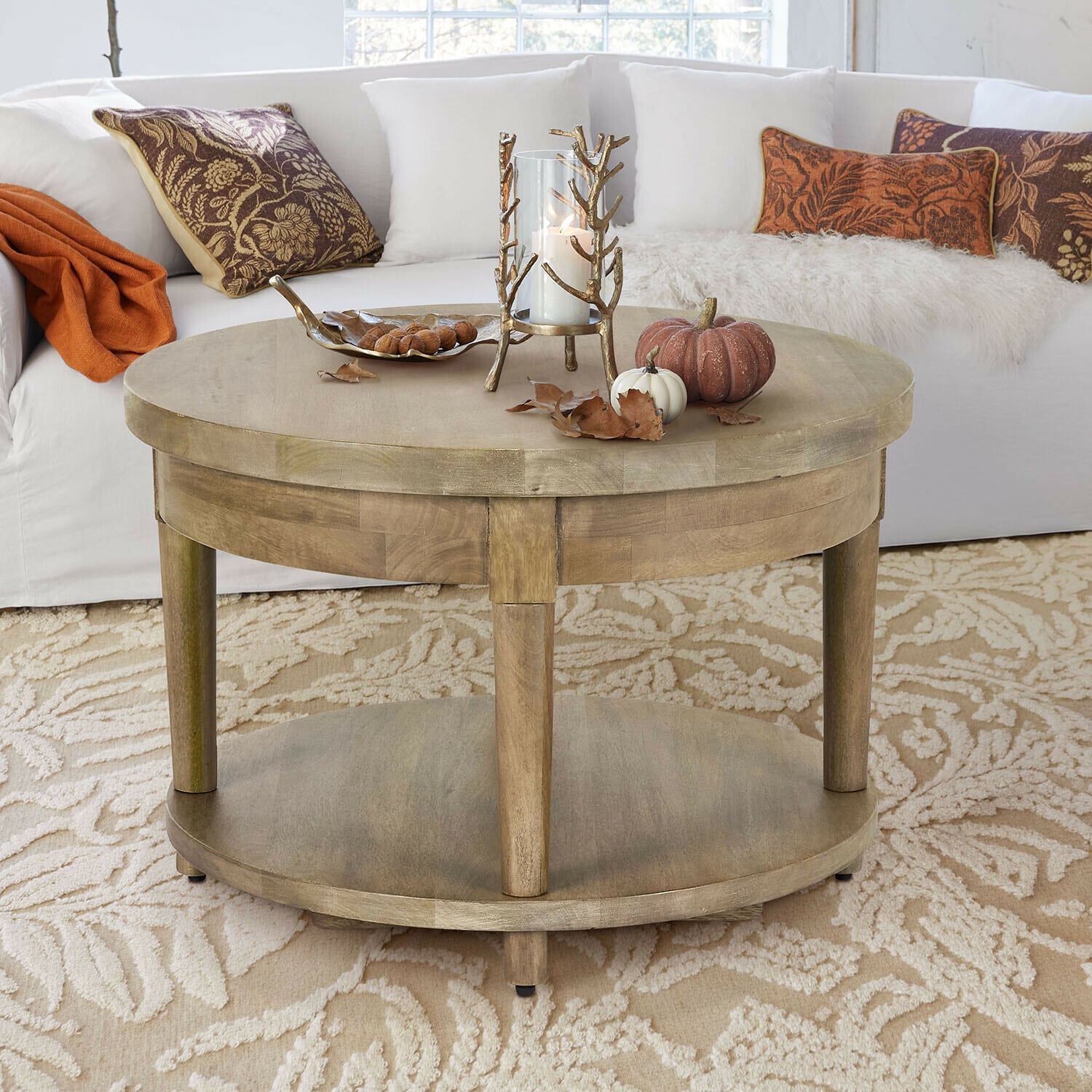 Mesa de centro Talinar Mesa de centro redonda de madera color natural con decoración de vela y calabazas sobre alfombra beige