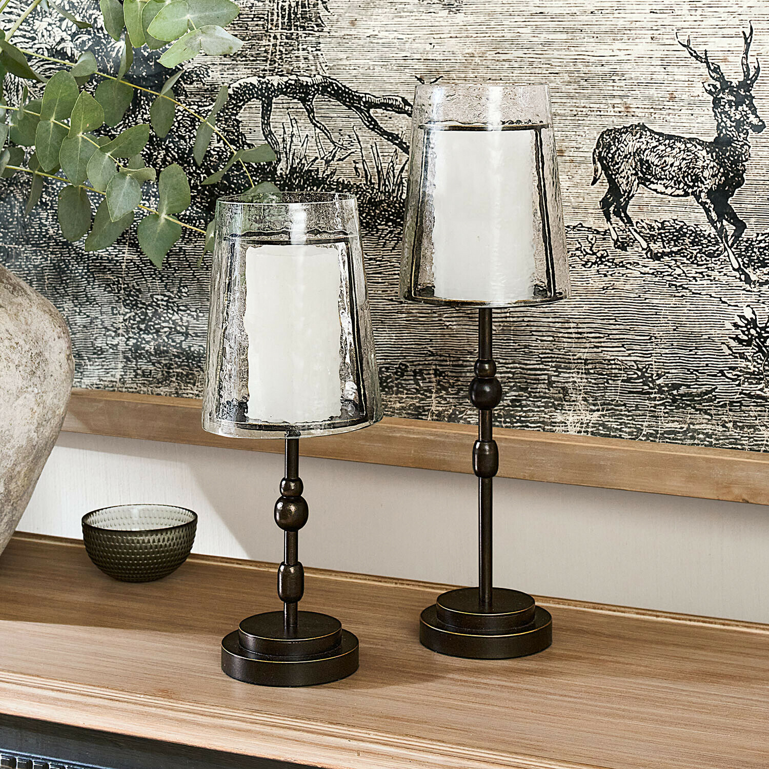 Dos candelabros de metal con pantallas de vidrio transparente y velas blancas sobre superficie de madera