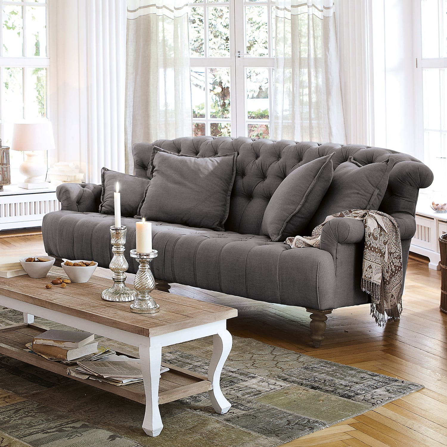 Sofá gris acolchado de tela con cojines y manta decorativa en salón luminoso con suelo de madera